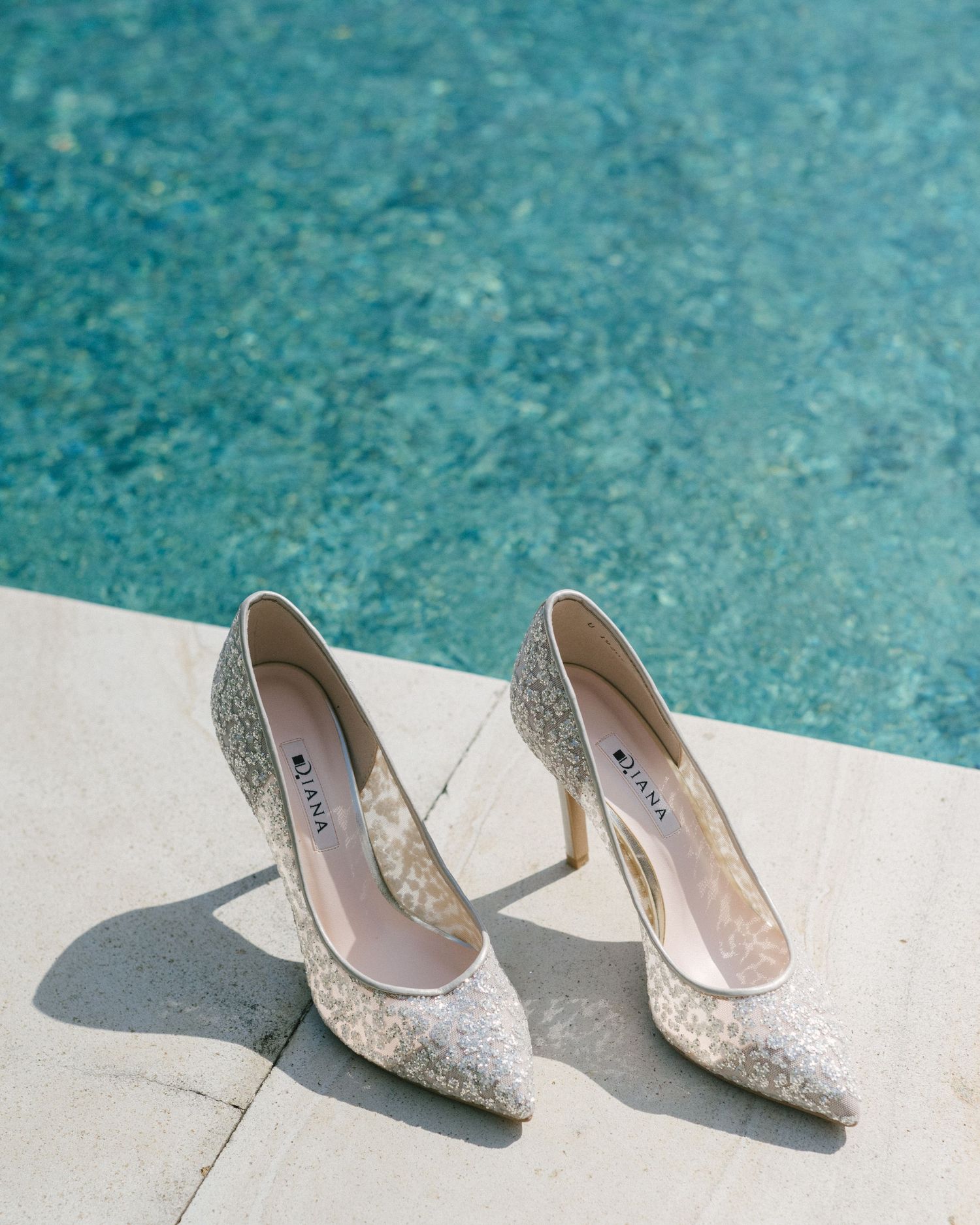 Sparkling glitter stiletto heels by LINX placed poolside on white marble near turquoise water.