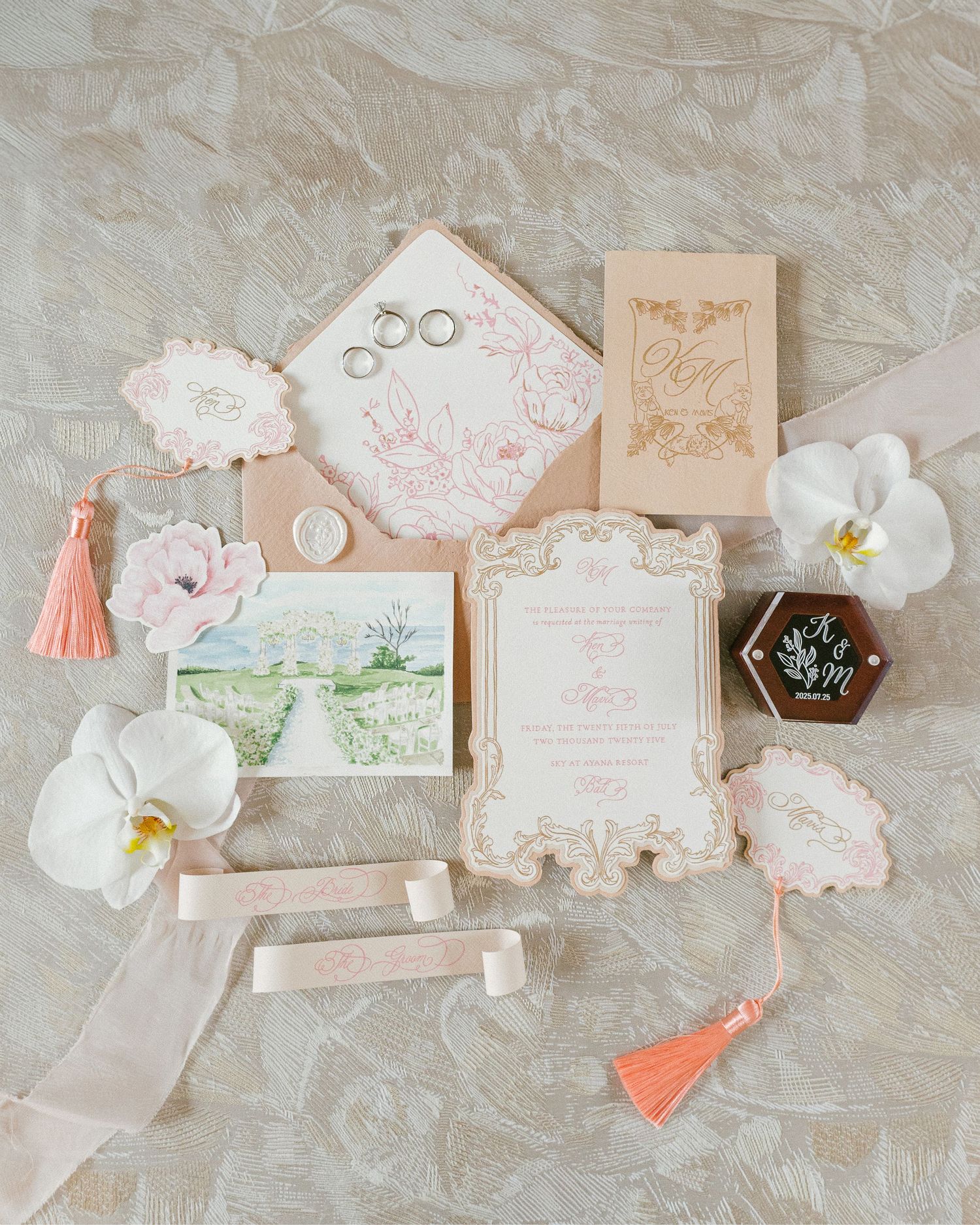 Elegant blush pink wedding invitation suite with floral details, tassel accents, white orchids, and gold rings on marble background.