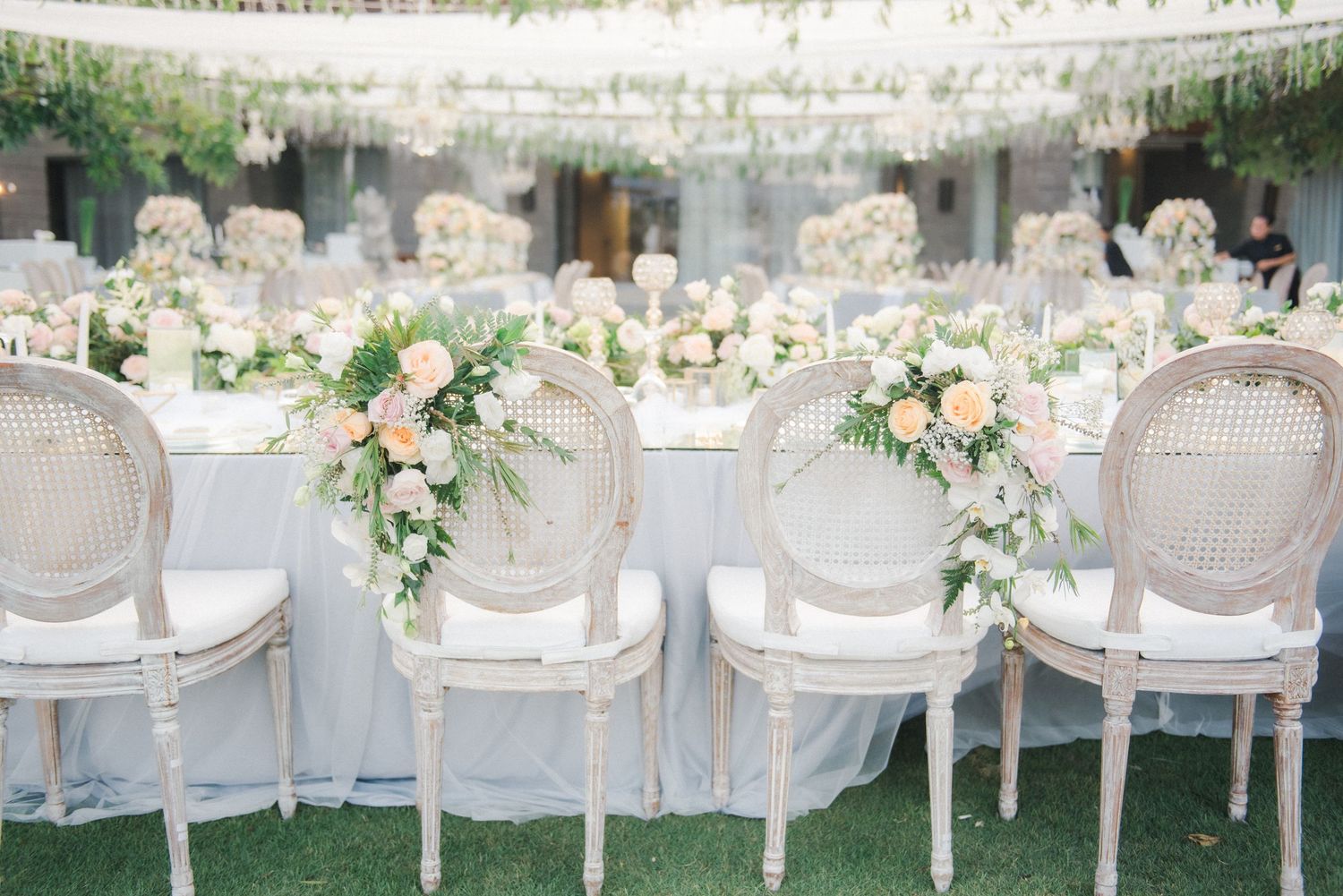 Elegant wedding reception with Louis XVI cane chairs adorned with blush floral arrangements at a white draped head table.