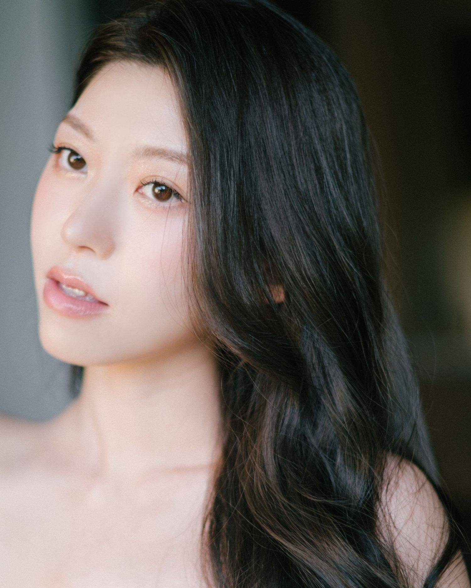 Young Asian woman with long dark wavy hair and natural makeup poses gracefully against a soft blurred background.