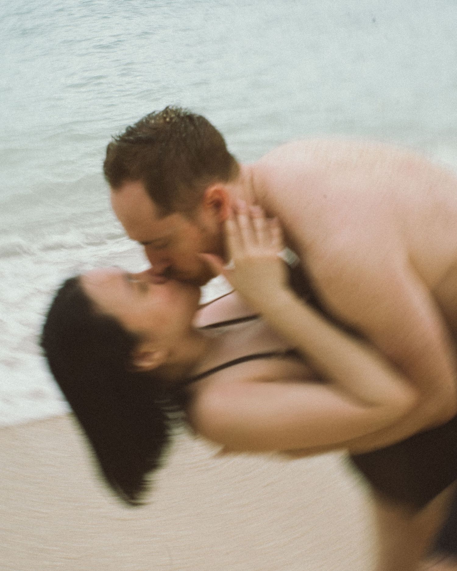 A couple shares a romantic kiss at the beach shoreline, with the man dipping the woman near the water.