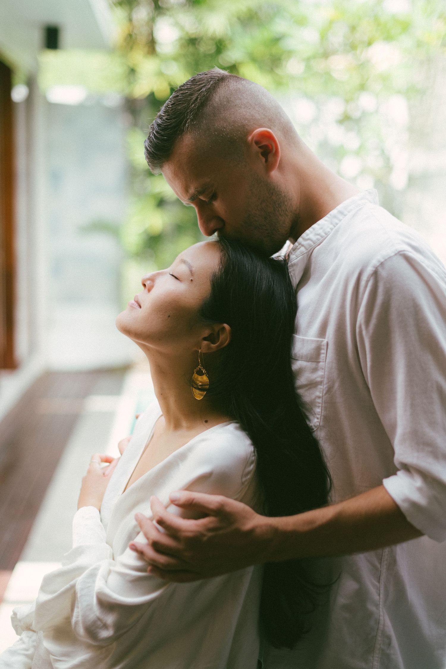 A couple shares a tender embrace, man kissing woman's forehead, both wearing white, with lush greenery in background.