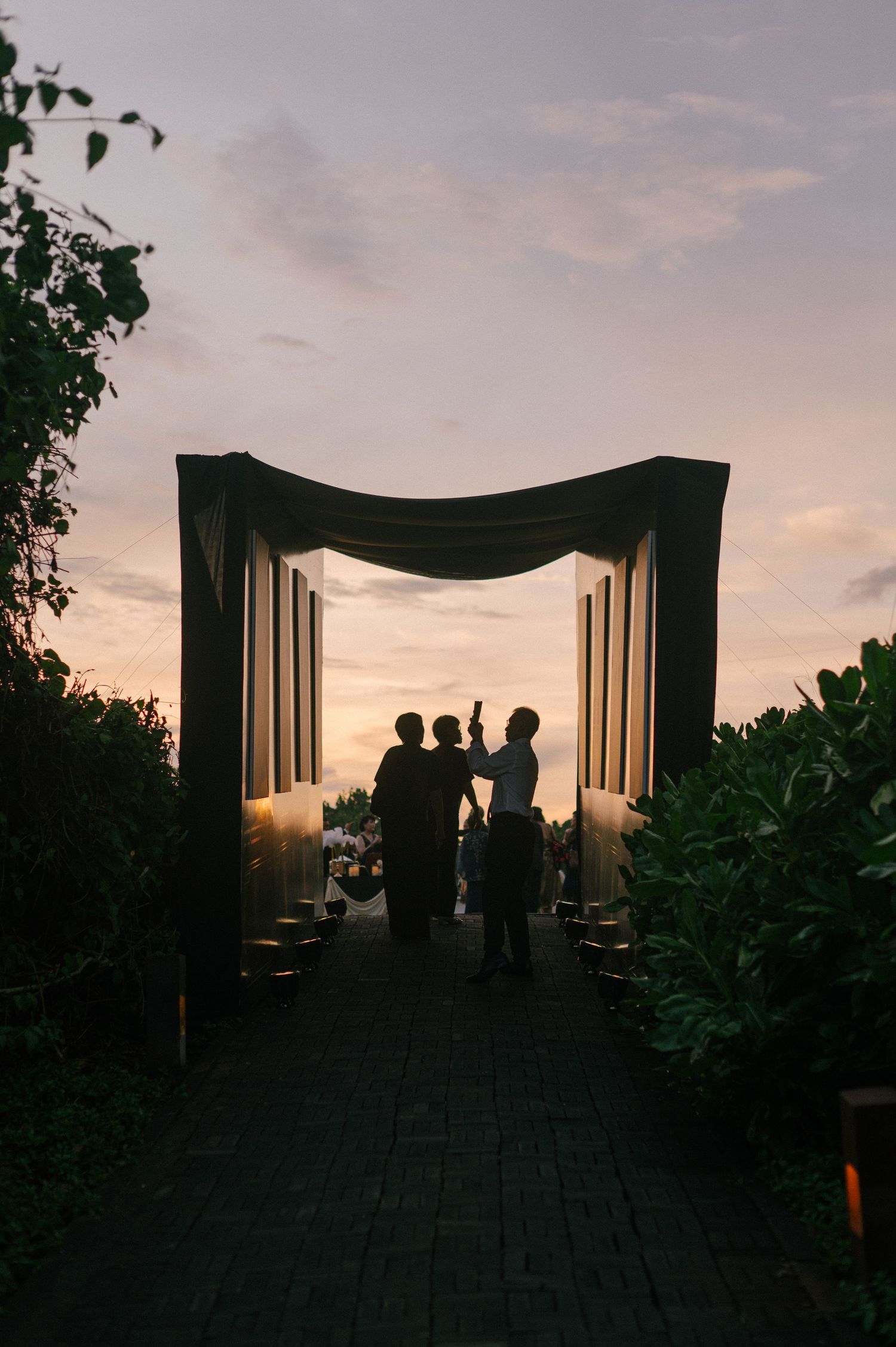 Silhouettes of people standing under a draped archway at sunset, surrounded by lush greenery and warm candlelight.