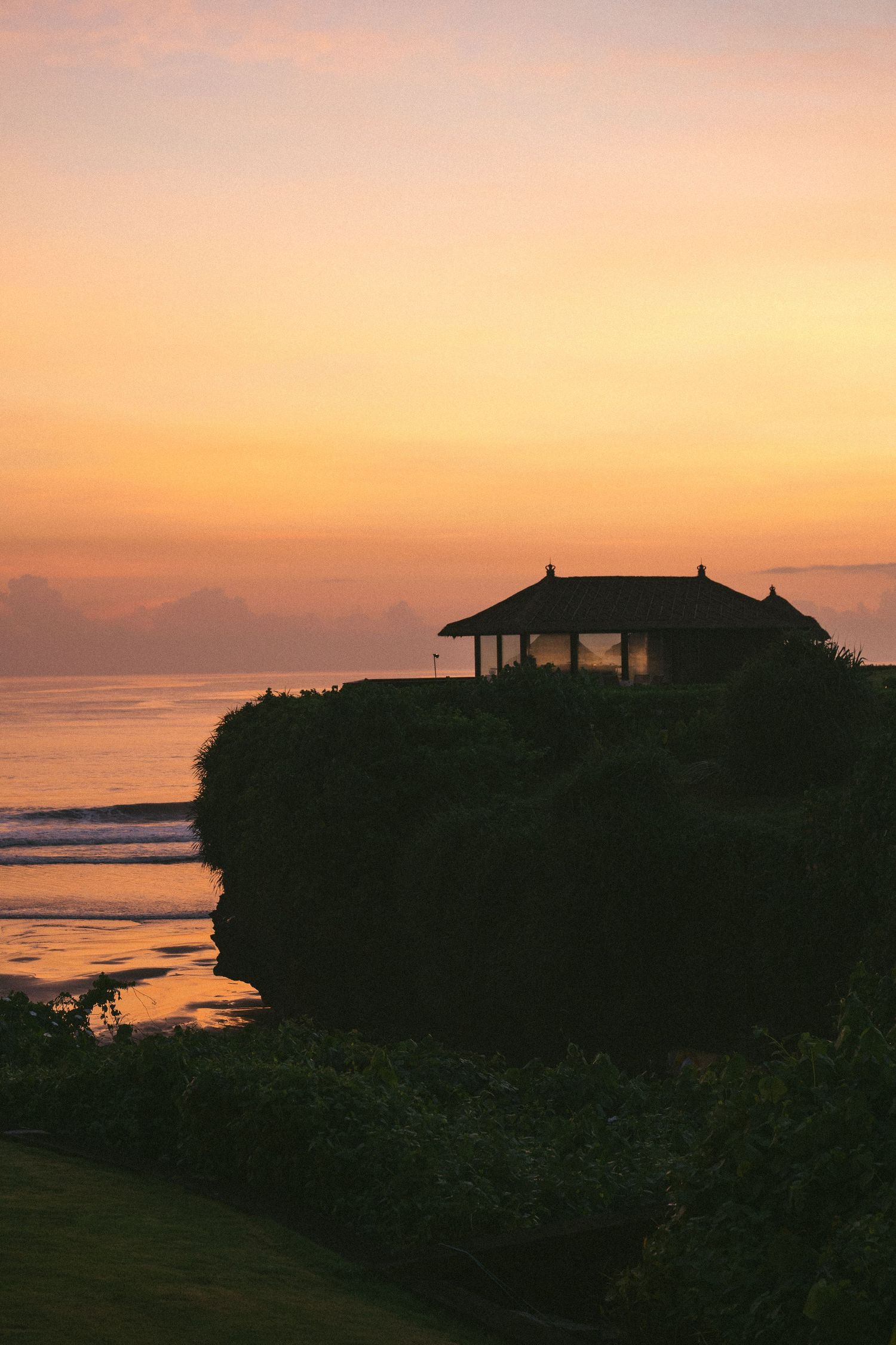 Silhouette of a Balinese temple on a rocky cliff at sunset with golden orange sky over the ocean.