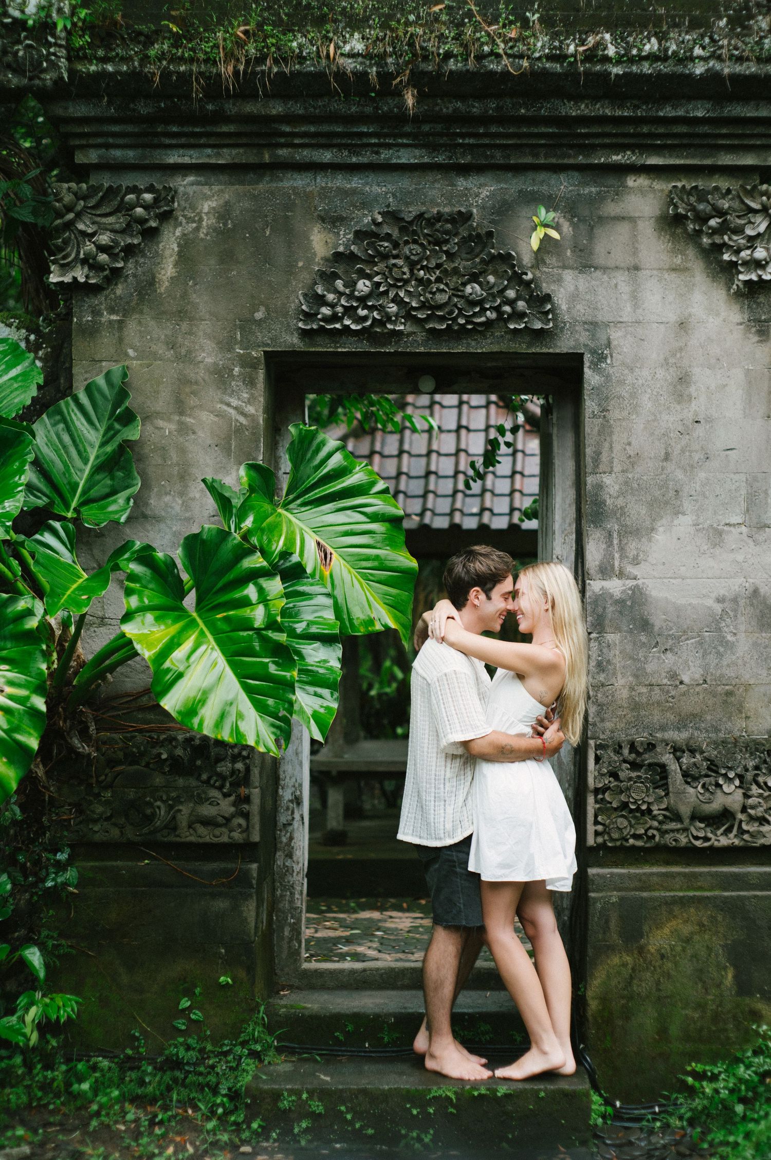 A couple shares a romantic kiss in front of an ornate Balinese stone temple doorway surrounded by lush tropical foliage.