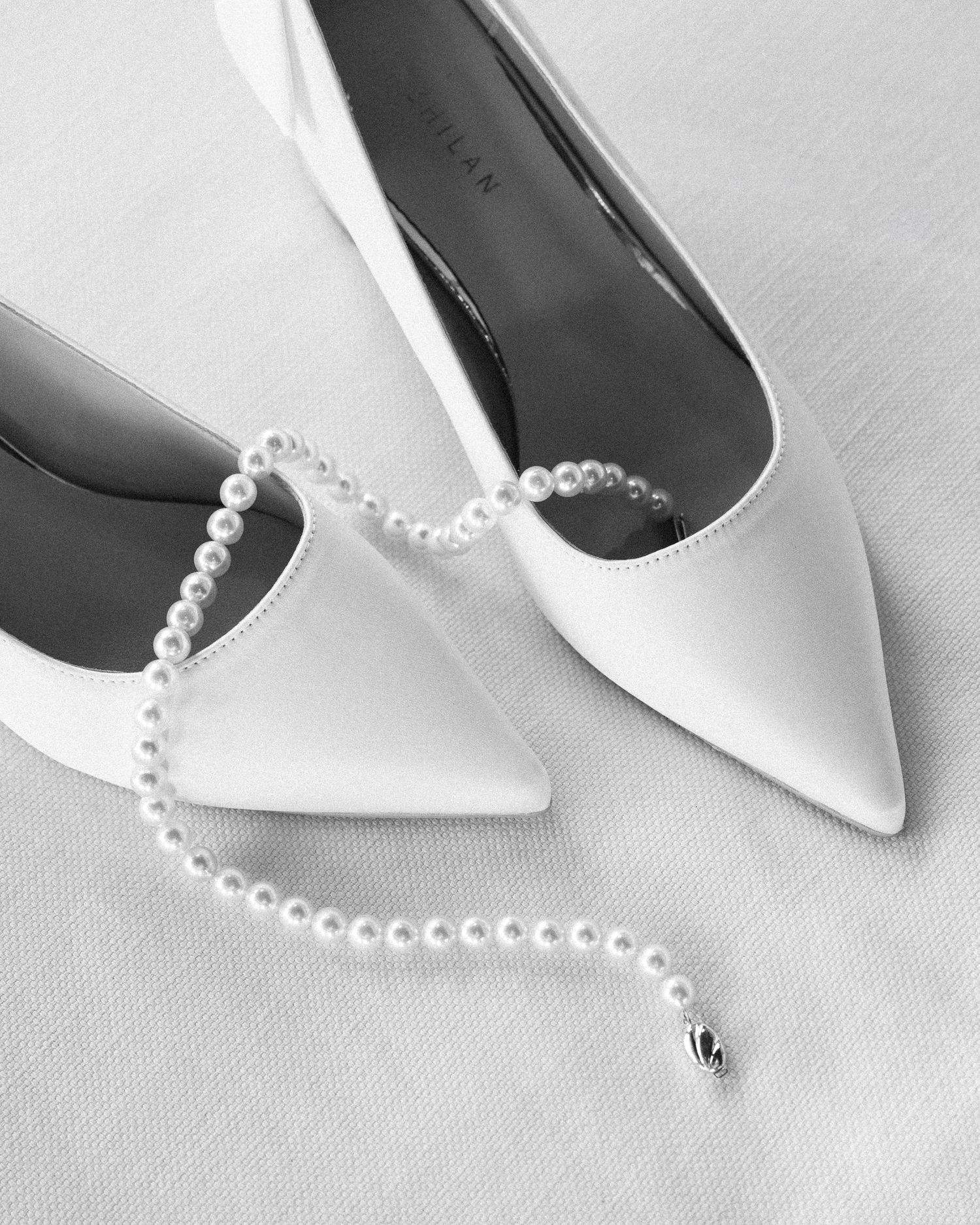 White satin pointed-toe heels with pearl strap accessory on white fabric, elegant bridal shoes in black and white photo.