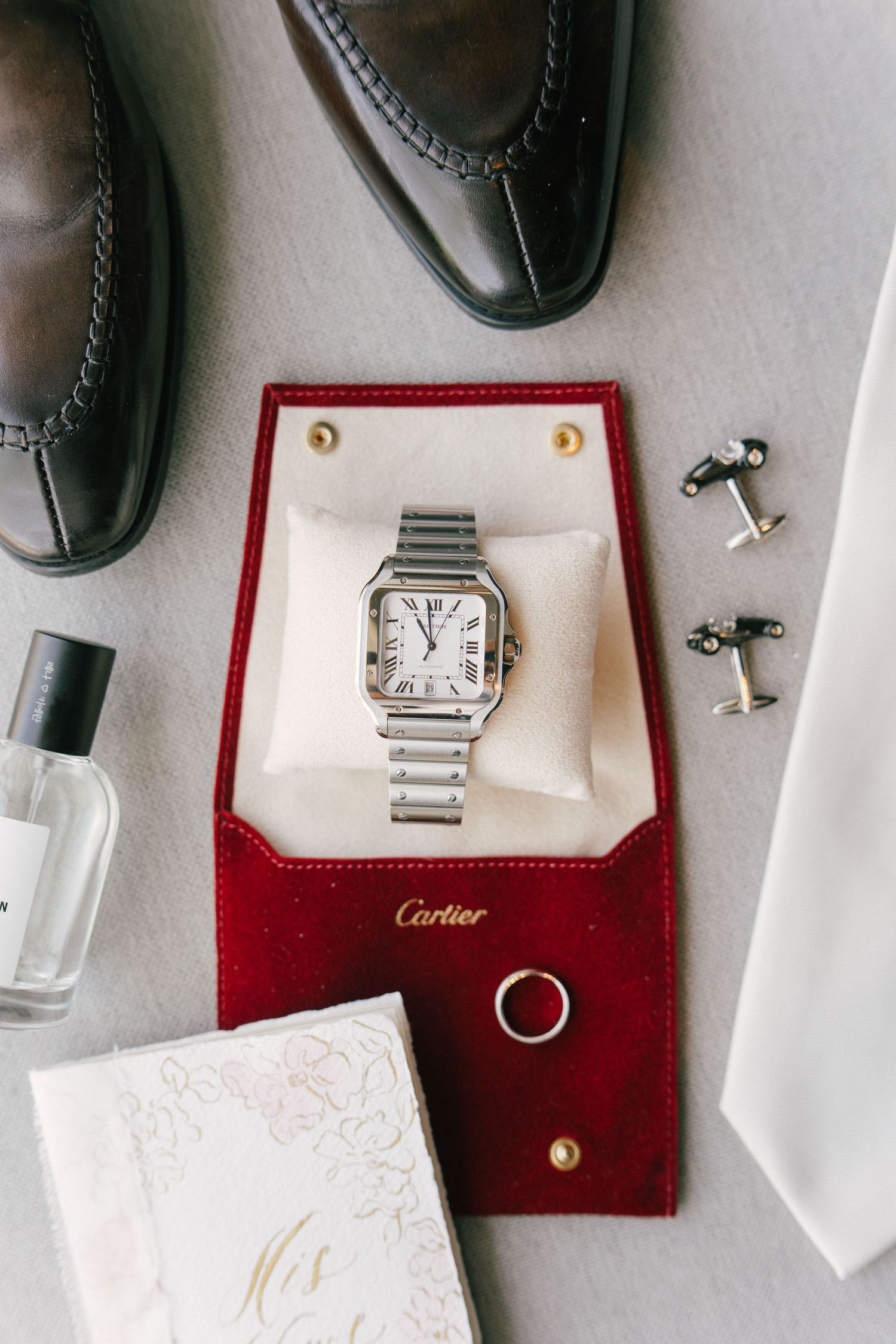 Cartier Santos watch displayed on red velvet case with wedding ring, cufflinks, dress shoes, cologne, and white tie flatlay.