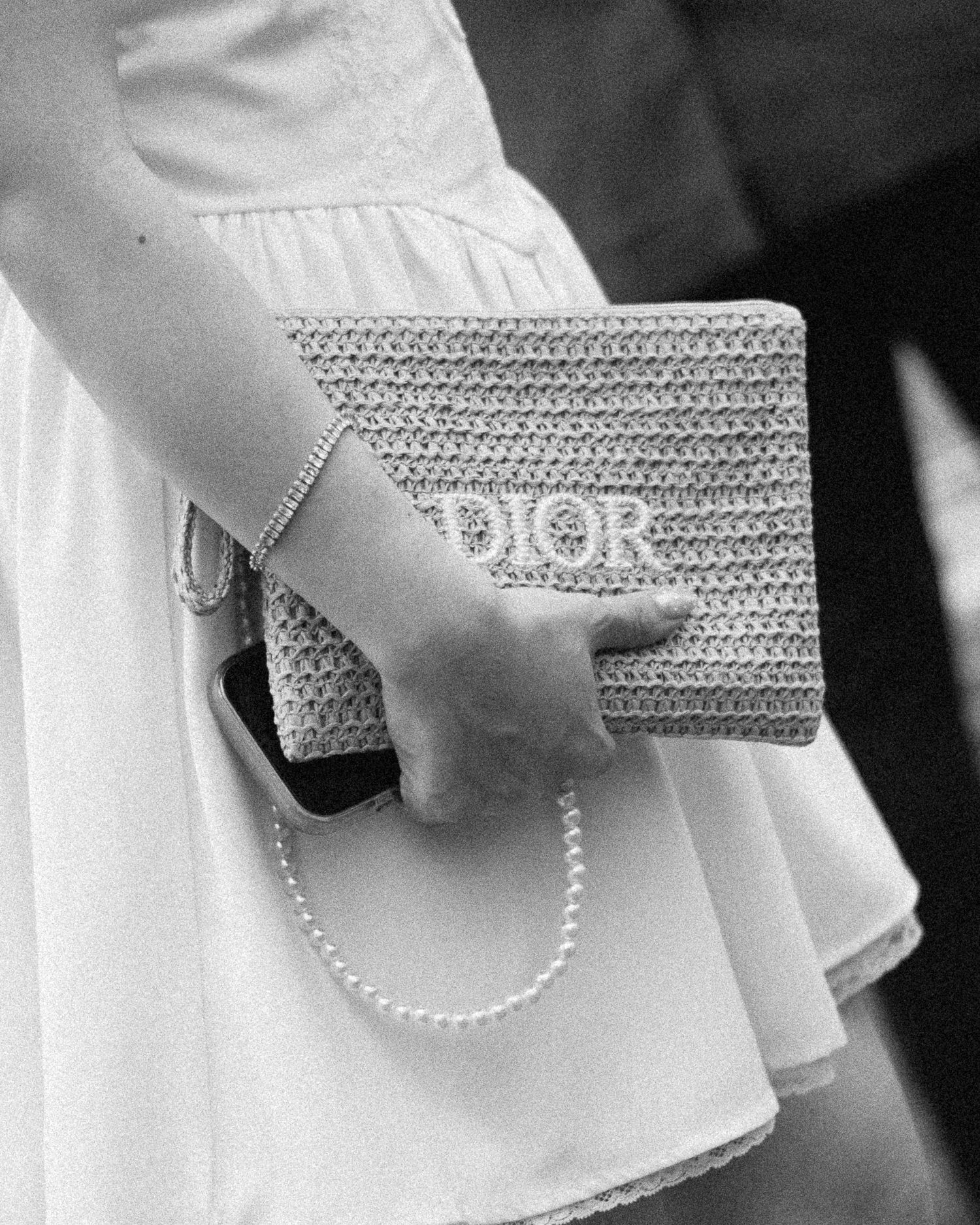 Black and white photo of a woman holding a woven Dior clutch bag, wearing a white dress and pearl bracelet.