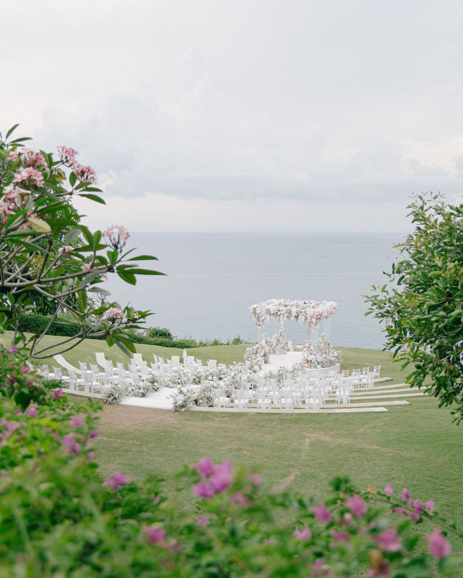 Elegant outdoor wedding ceremony setup with white chairs and floral arch overlooking the ocean, framed by tropical blooms.