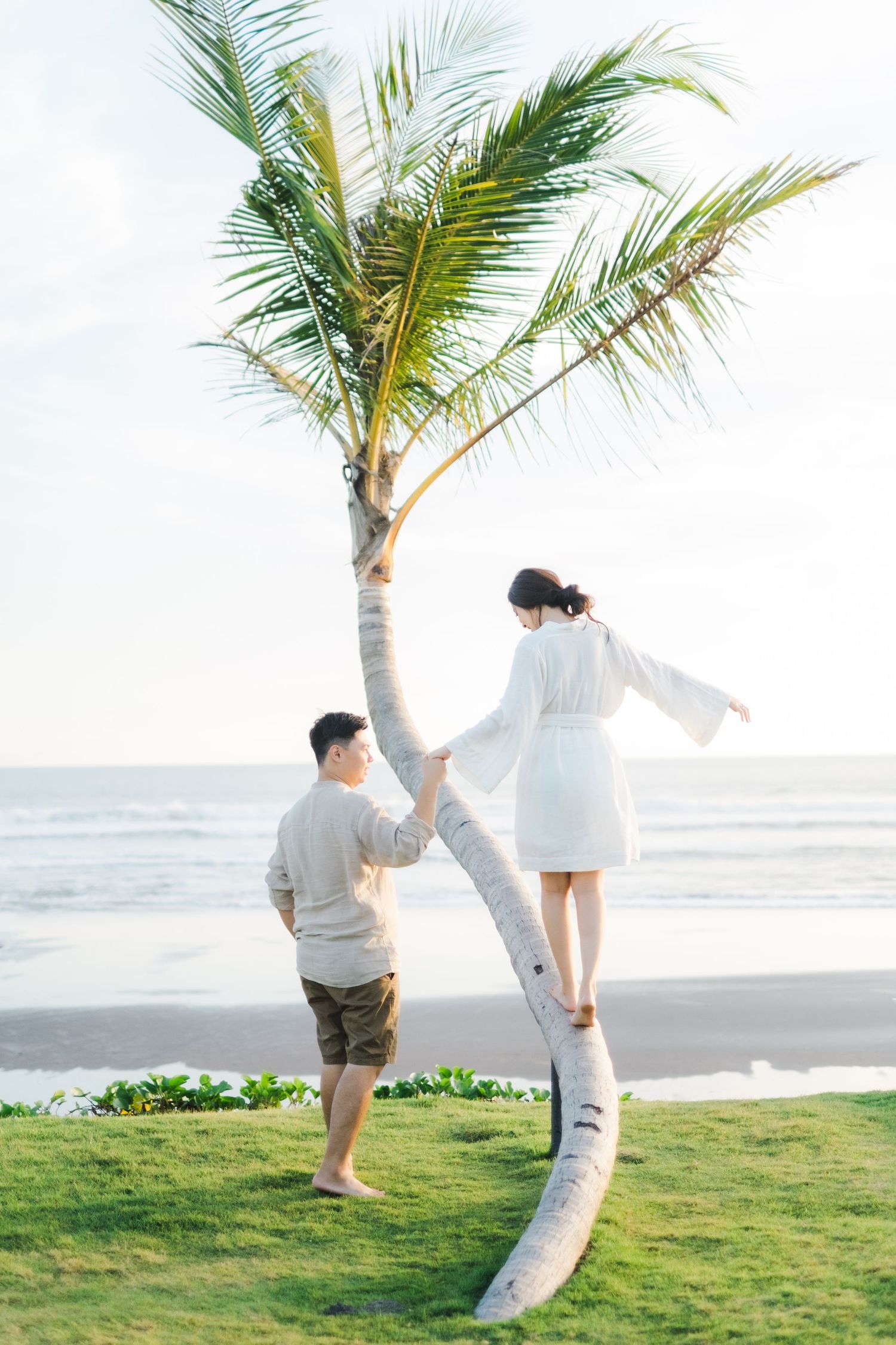 Couple posing by a leaning palm tree on a tropical beach, woman in white dress balancing on trunk, ocean in background.