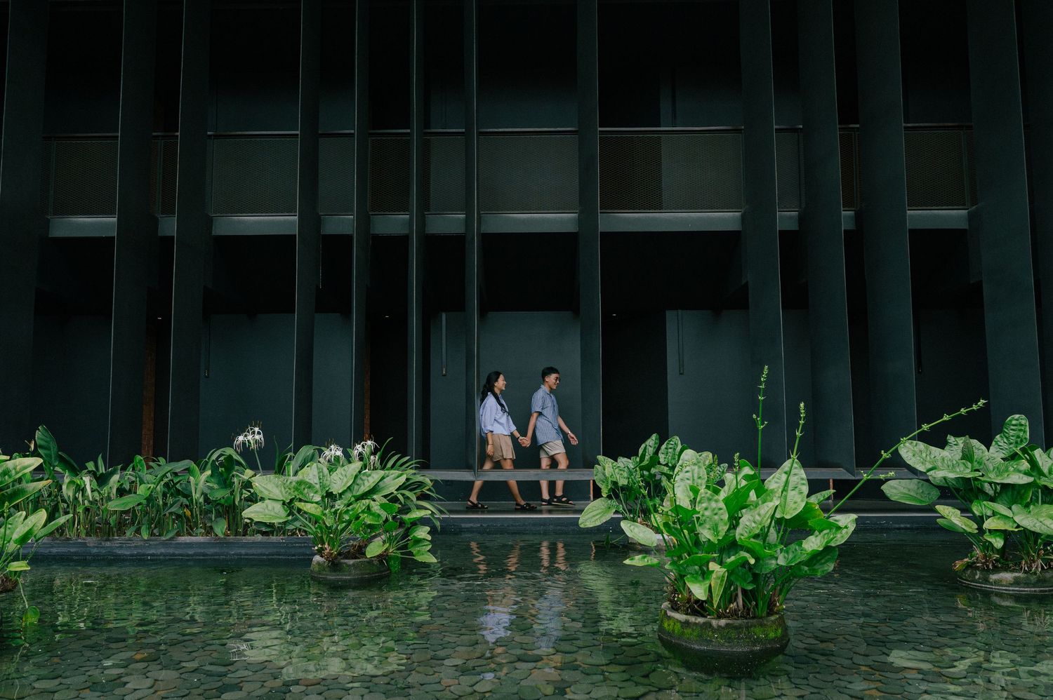 Couple walking hand in hand through a lush tropical garden with green plants and a modern dark building backdrop.