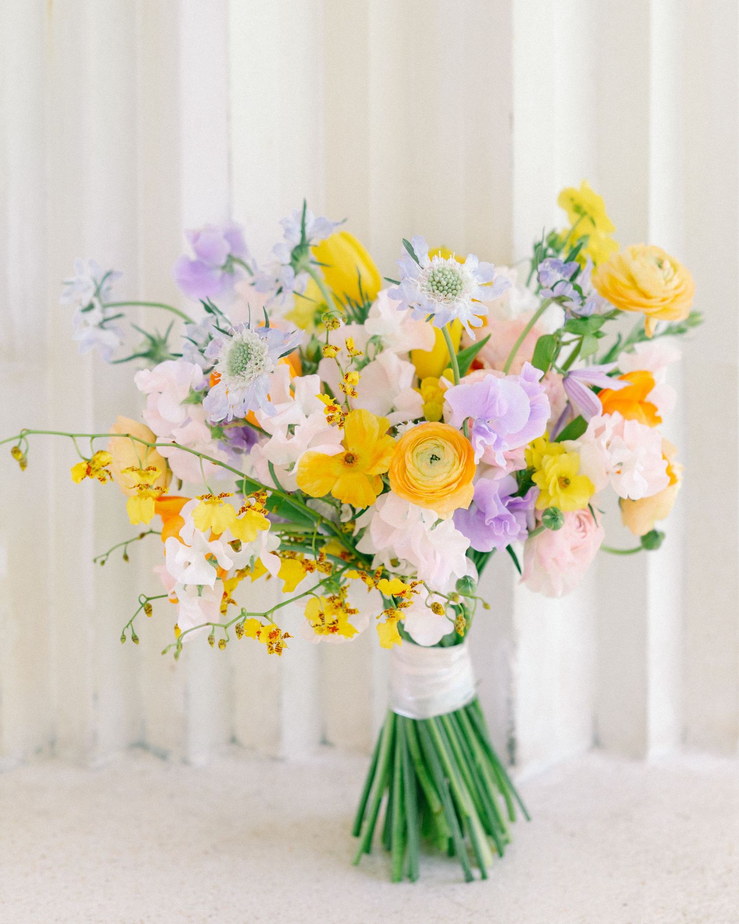 Vibrant spring wedding bouquet featuring yellow ranunculus, lavender lisianthus, pink blooms, and greenery wrapped in white ribbon.