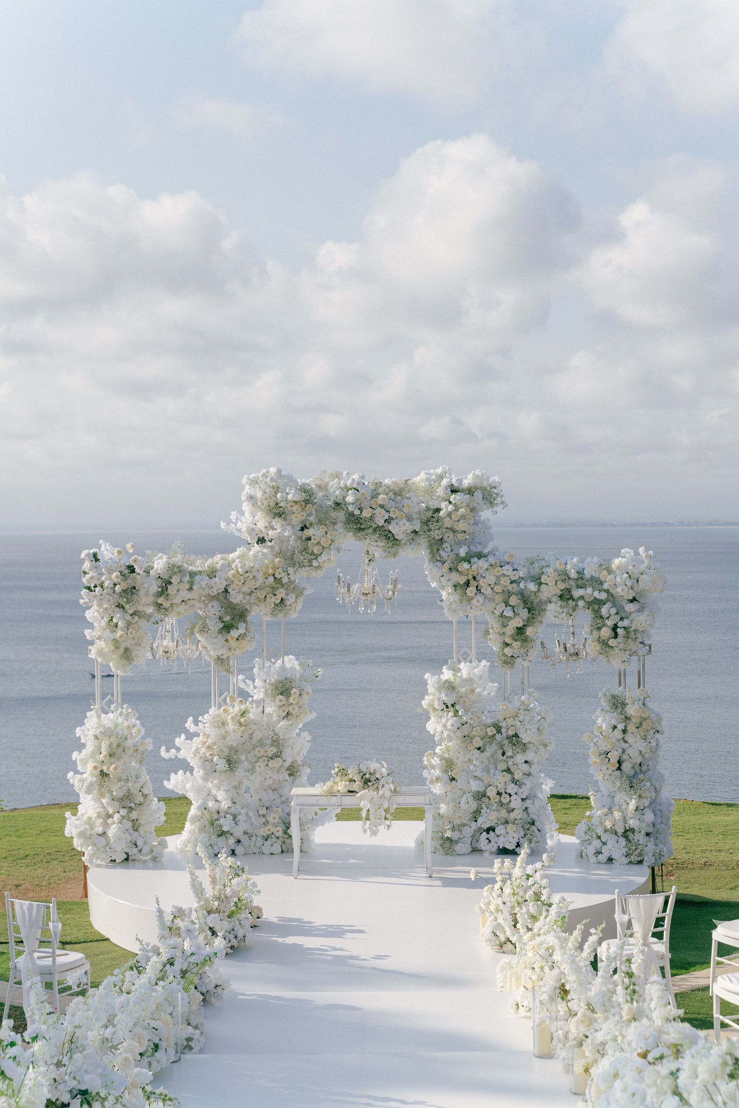 Elegant all-white outdoor wedding ceremony arch adorned with lush florals, chandelier, and ocean backdrop.