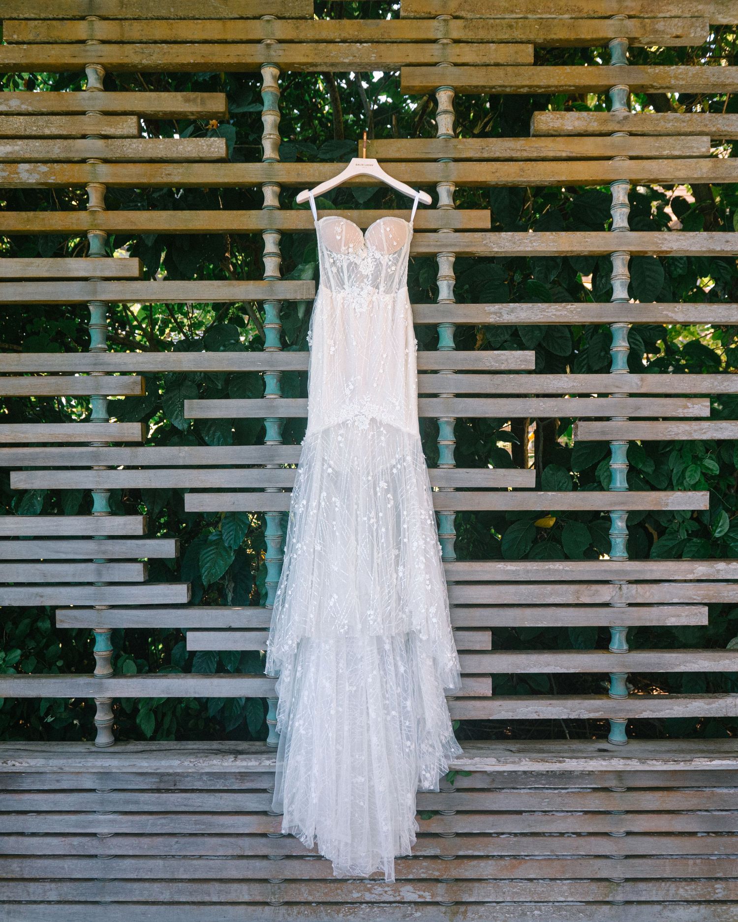 Elegant strapless lace wedding gown with tiered skirt hanging on a wooden lattice backdrop surrounded by greenery.