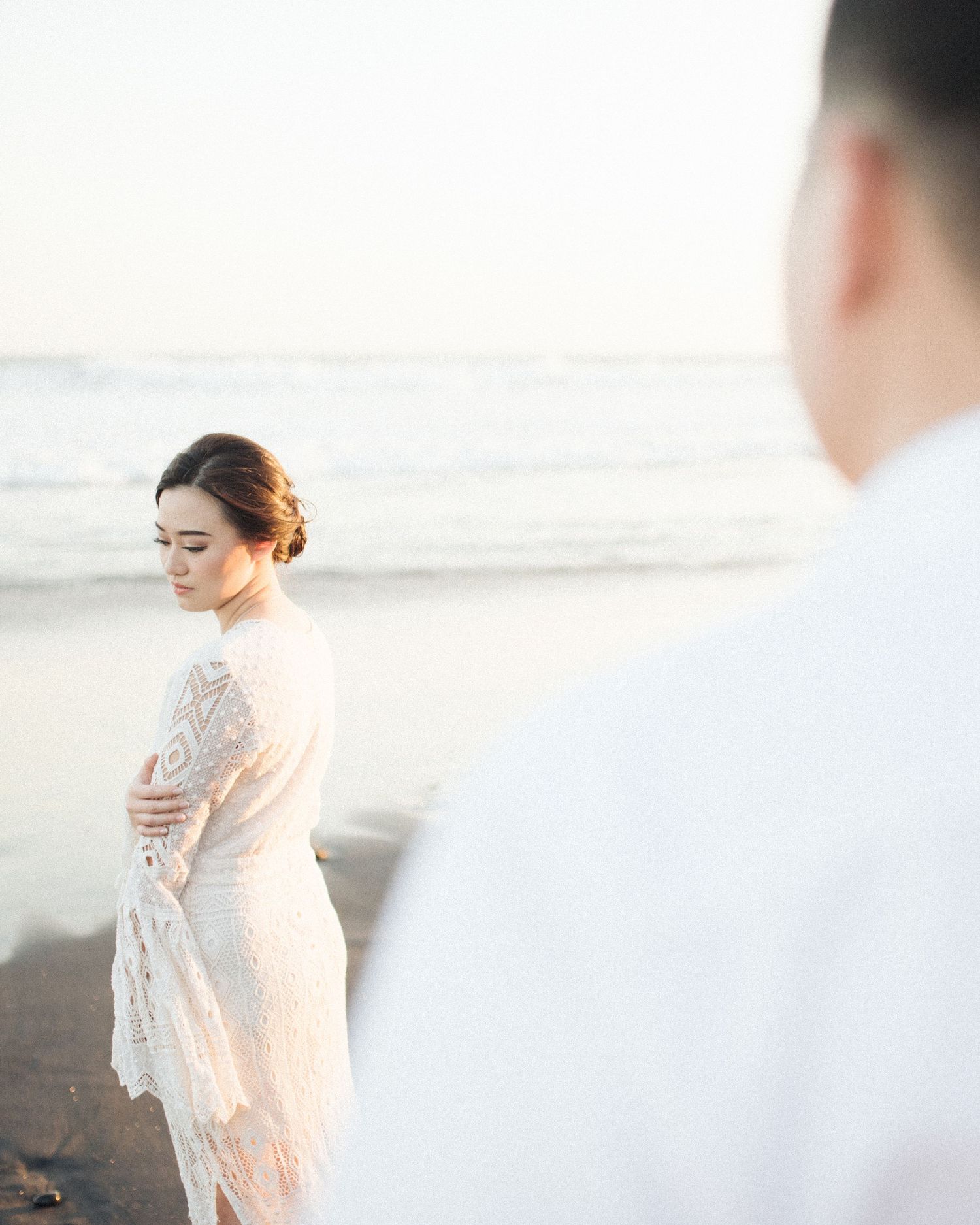 A bride in a white lace dress stands on a beach at sunset, with a groom in white blurred in the foreground.