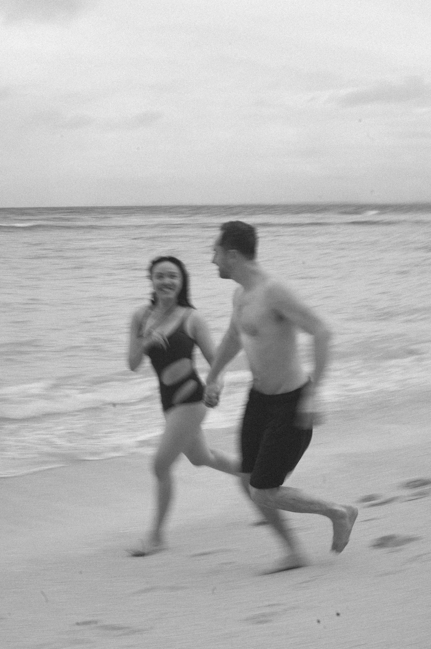 Black and white photo of a smiling couple holding hands and running together on a sandy beach near the ocean.
