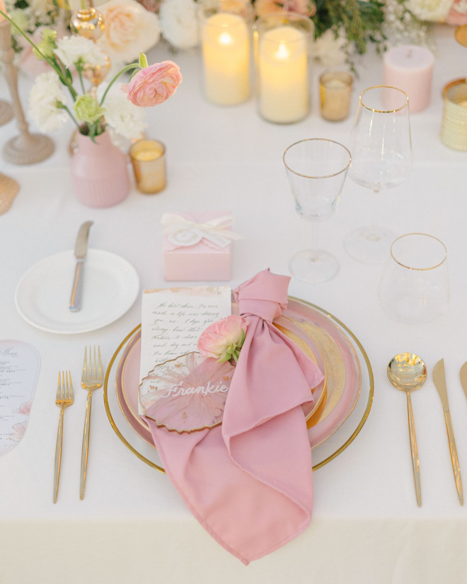 Elegant pink and gold wedding table setting with floral napkin, gold cutlery, candles, and blush flowers.