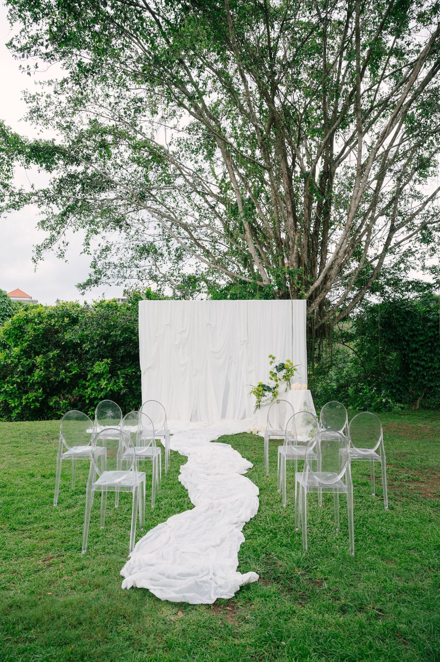 Elegant outdoor wedding ceremony setup with white drape backdrop, ghost chairs, and flowing aisle runner on lush green lawn.