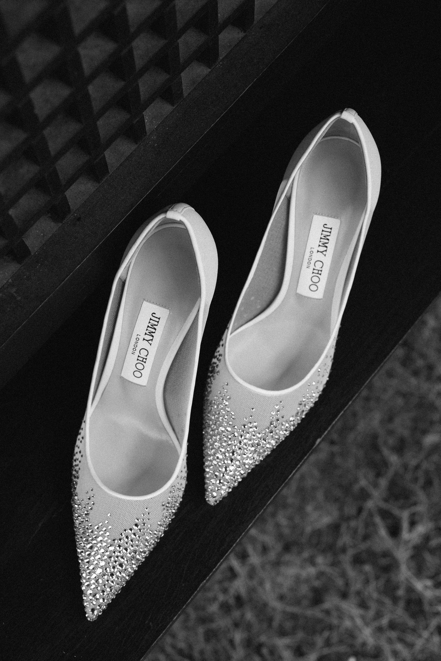 Black and white photo of crystal-embellished Jimmy Choo pointed-toe heels displayed on a dark surface.