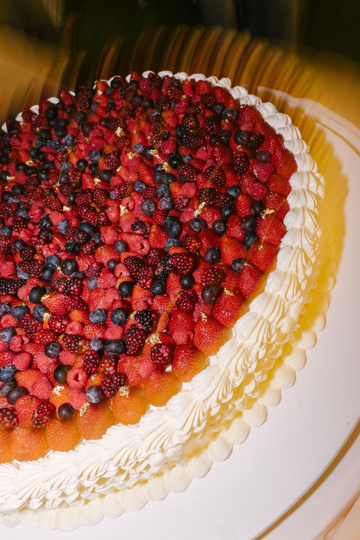 Elegant white cake topped with fresh mixed berries including raspberries, blueberries, and blackberries with gold leaf accents.