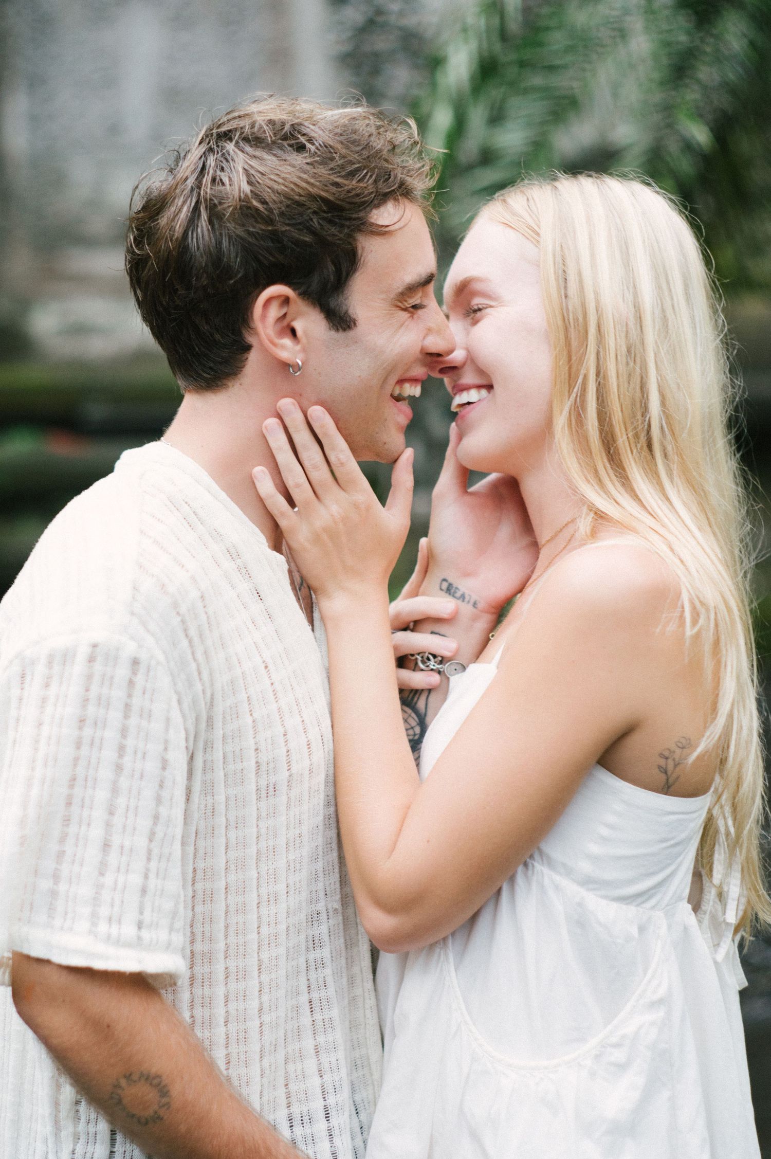 A joyful couple in white outfits share a romantic moment, smiling and almost kissing outdoors.