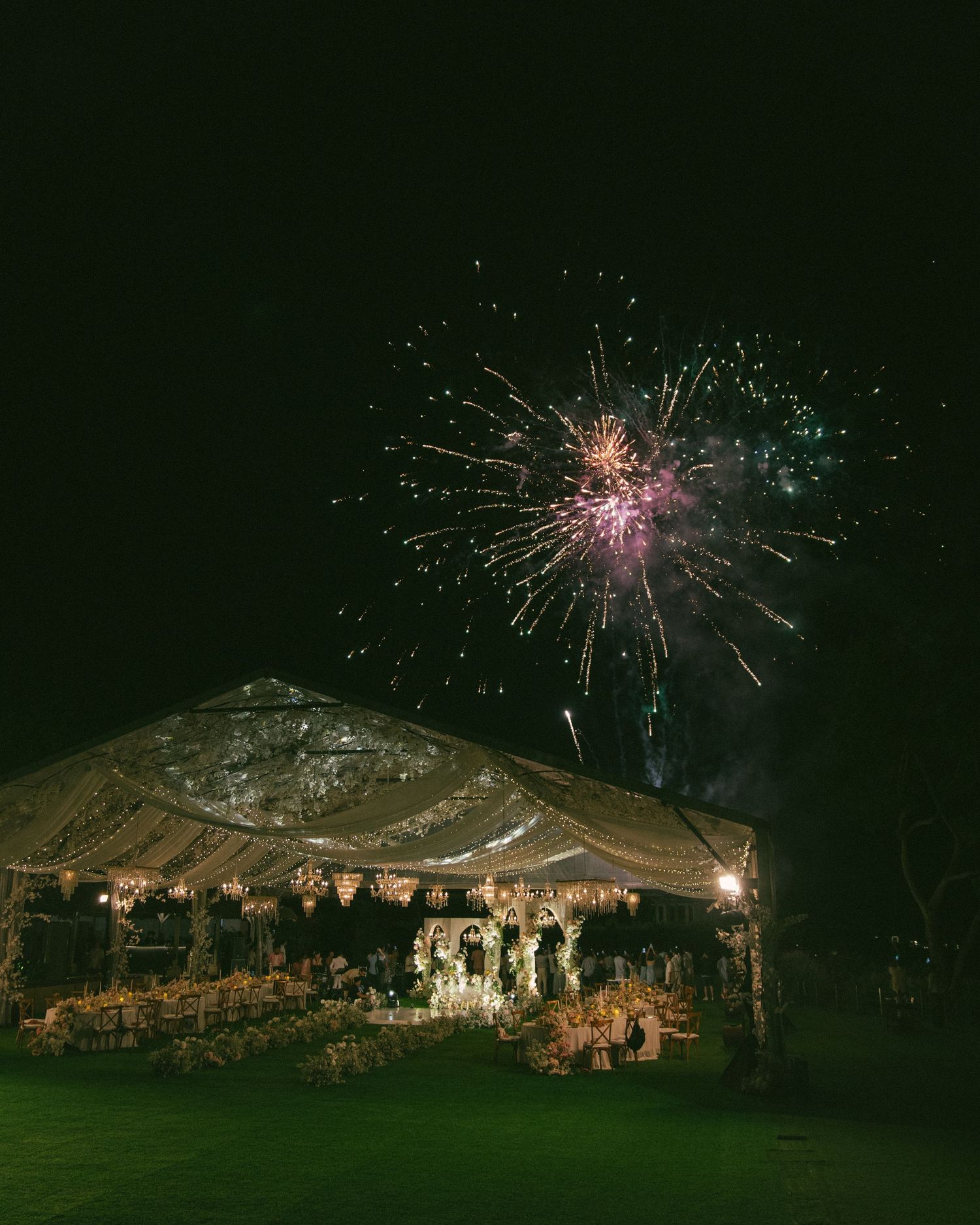 Elegant outdoor wedding tent illuminated at night with colorful fireworks bursting in the dark sky above.