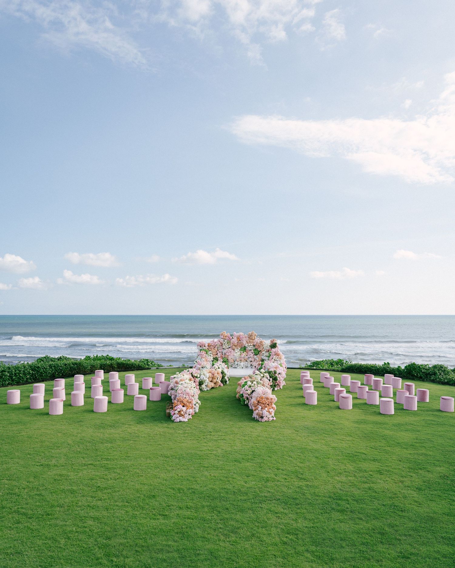 Outdoor beach wedding ceremony setup with pink chairs, floral arch, and ocean backdrop on lush green lawn.