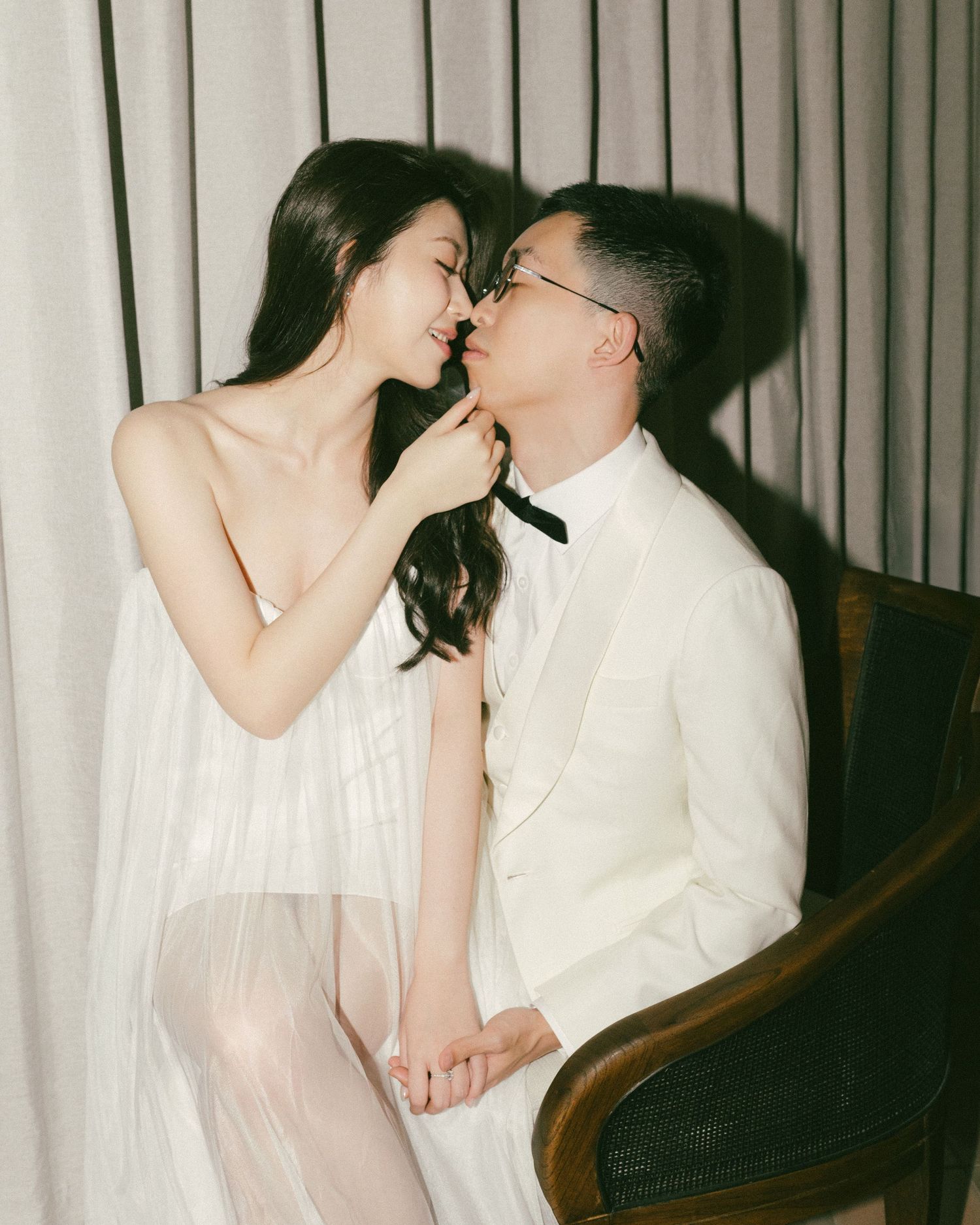 A couple in elegant white wedding attire share a romantic moment, with the woman in a sheer gown and man in a white tuxedo.