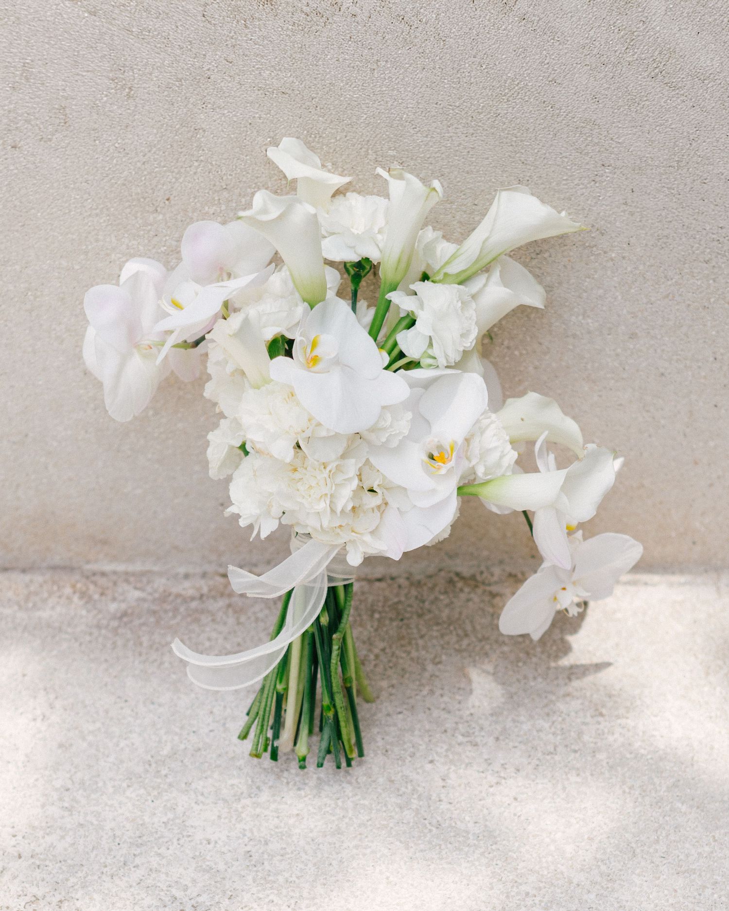 Elegant white wedding bouquet featuring calla lilies, orchids, and hydrangeas tied with ribbon, displayed against a neutral wall.