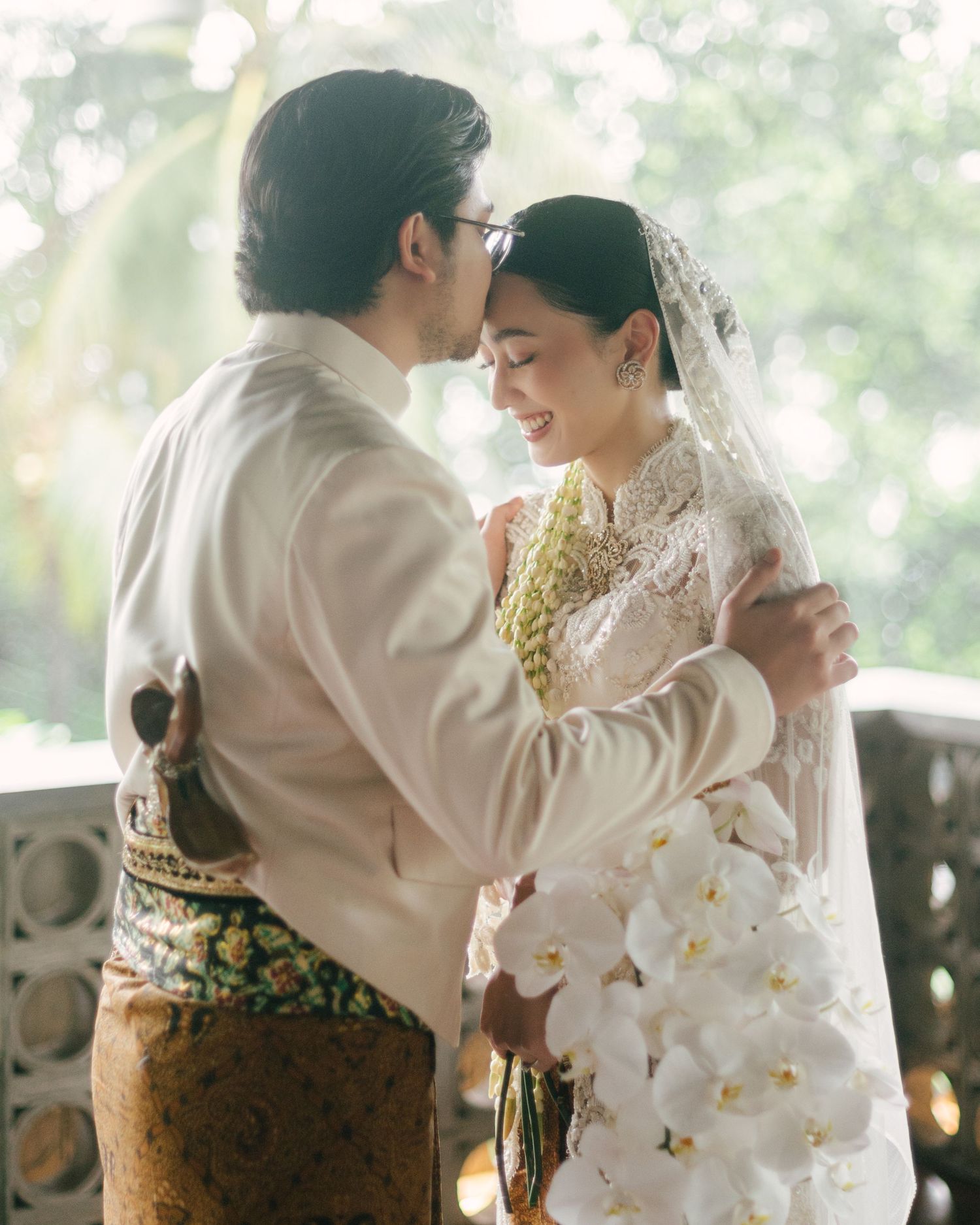 Couple in traditional Balinese wedding attire sharing an intimate moment, bride holding white orchid bouquet with lace veil.