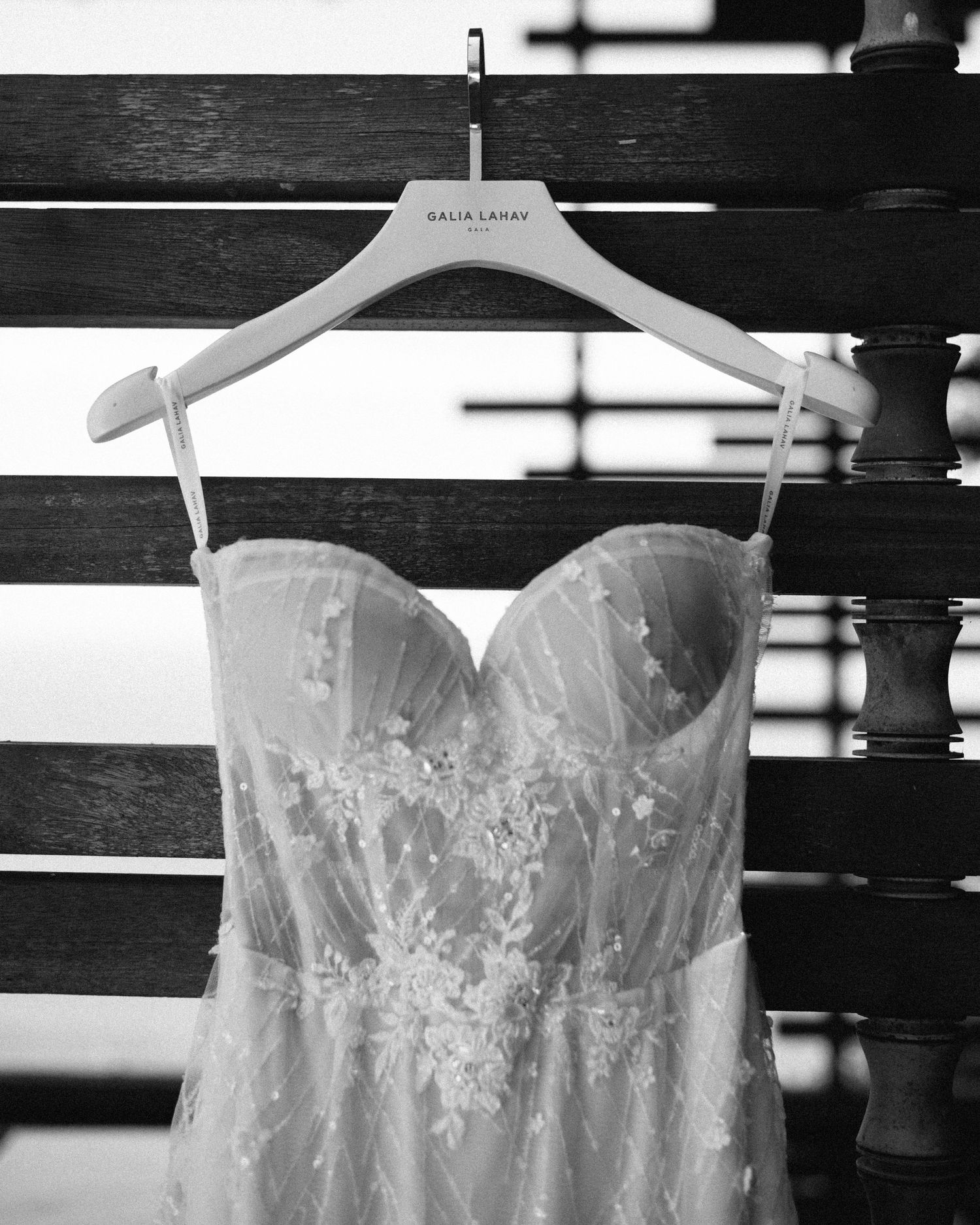 Black and white photo of a Galia Lahav lace wedding dress hanging on a branded hanger against wooden slats.