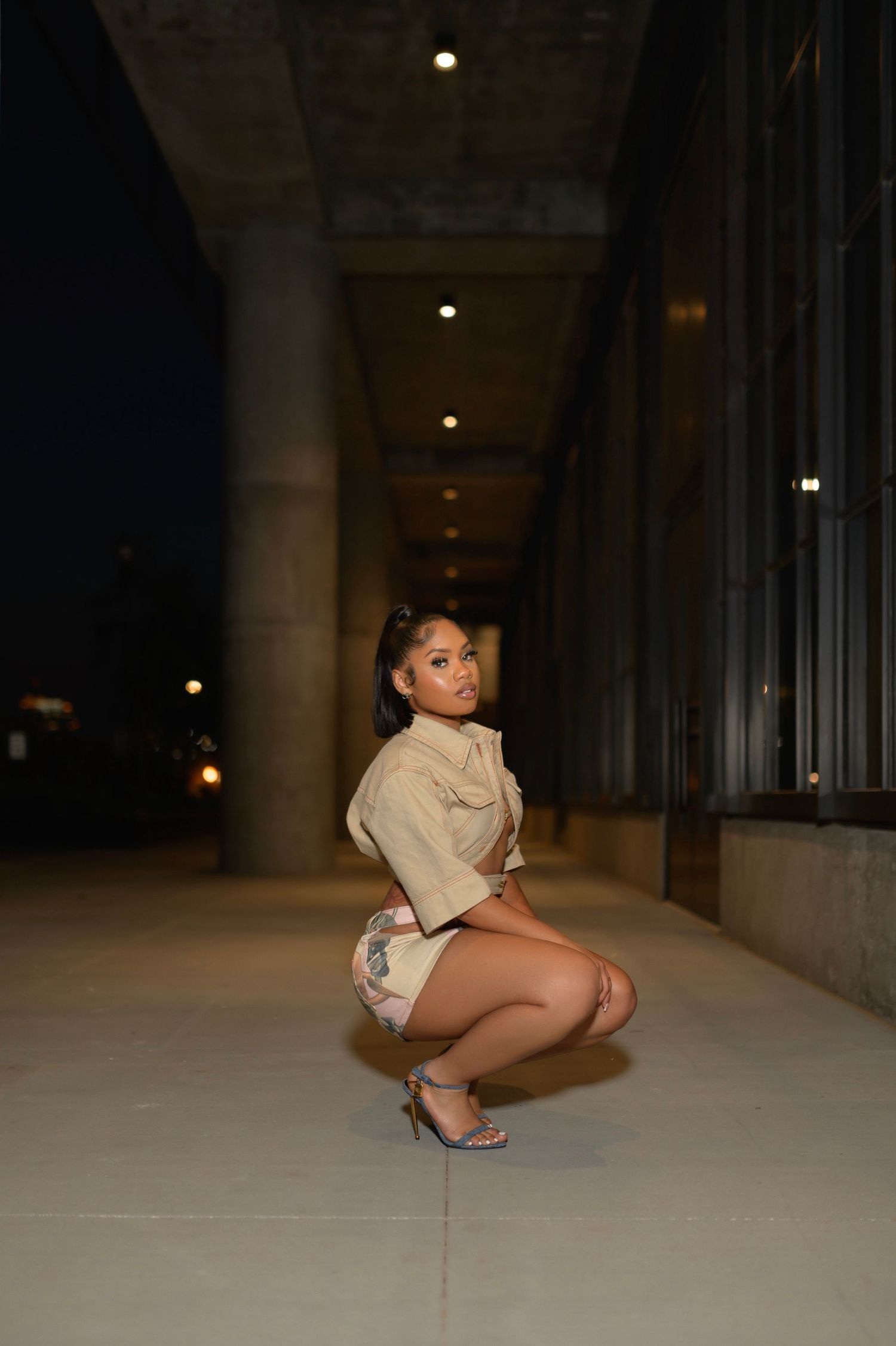 Woman in beige crop jacket and floral shorts squatting in a nighttime outdoor corridor with heels.