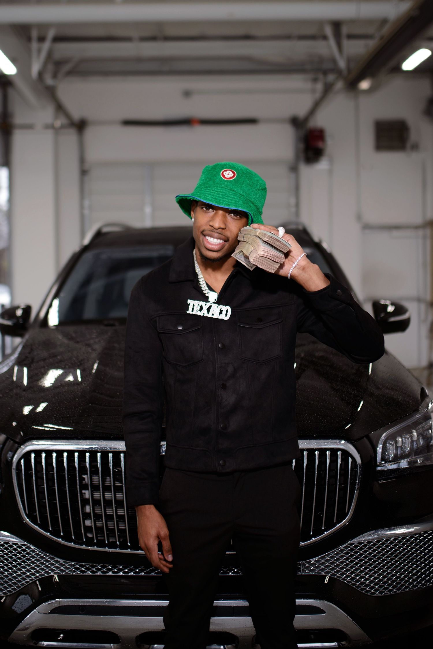 Man in black outfit and green bucket hat poses in front of a black luxury SUV in a garage.