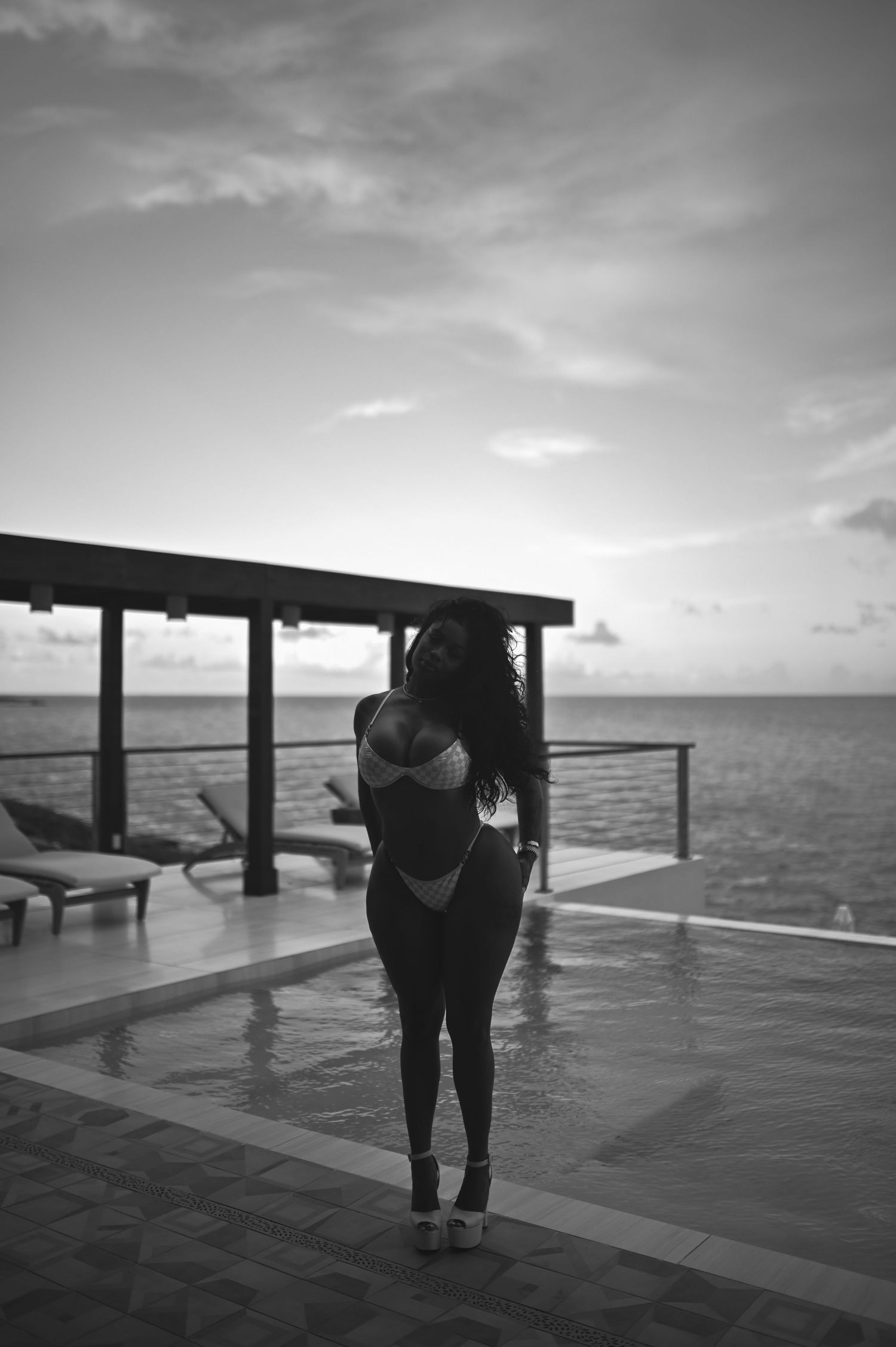 Black and white photo of a woman in a bikini posing by an infinity pool overlooking the ocean at sunset.