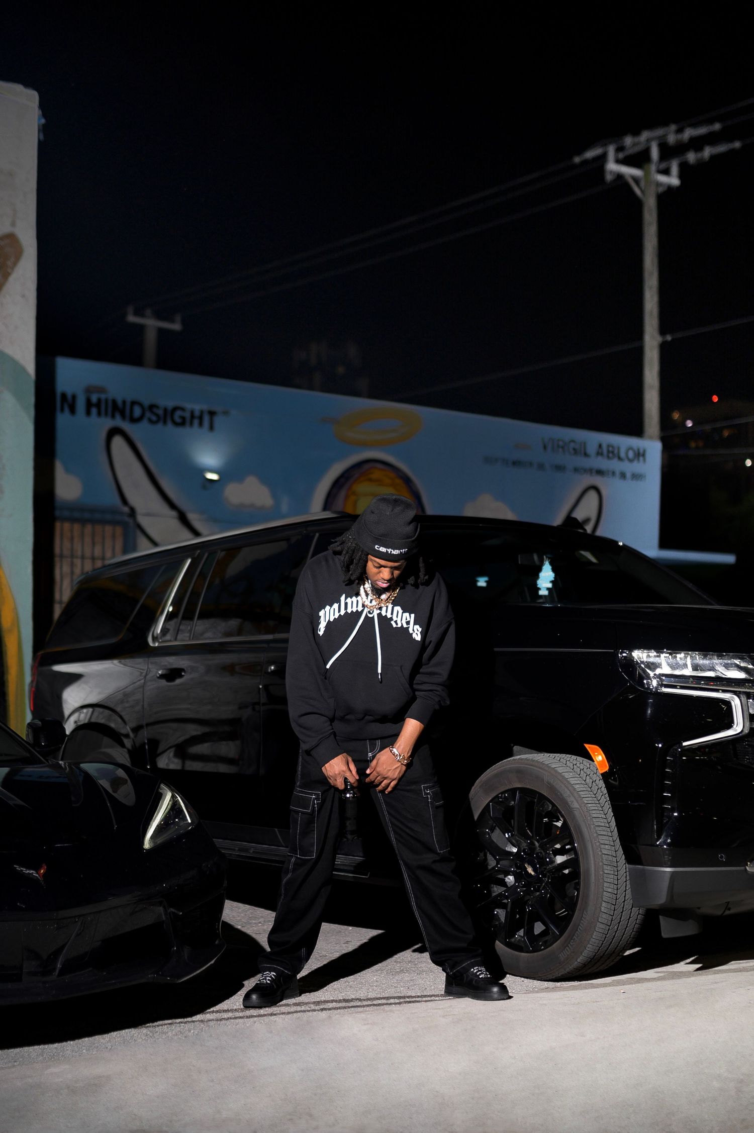 Man in black Palm Angels hoodie leaning against a black SUV at night outside an art gallery.