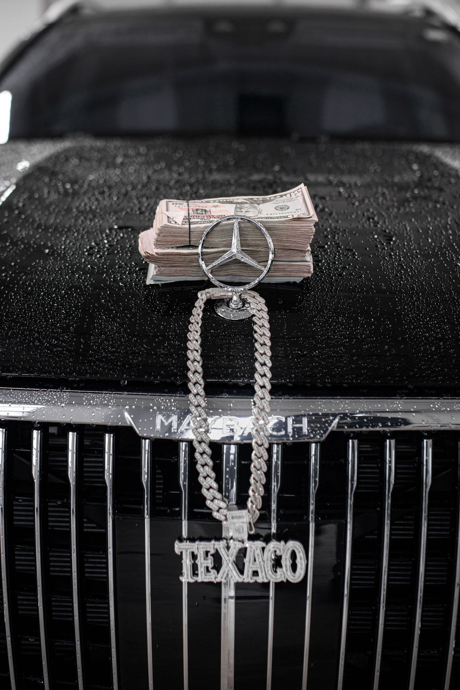 Stack of cash bills on a wet black Mercedes-Maybach hood with a silver Cuban link chain and Texaco pendant.