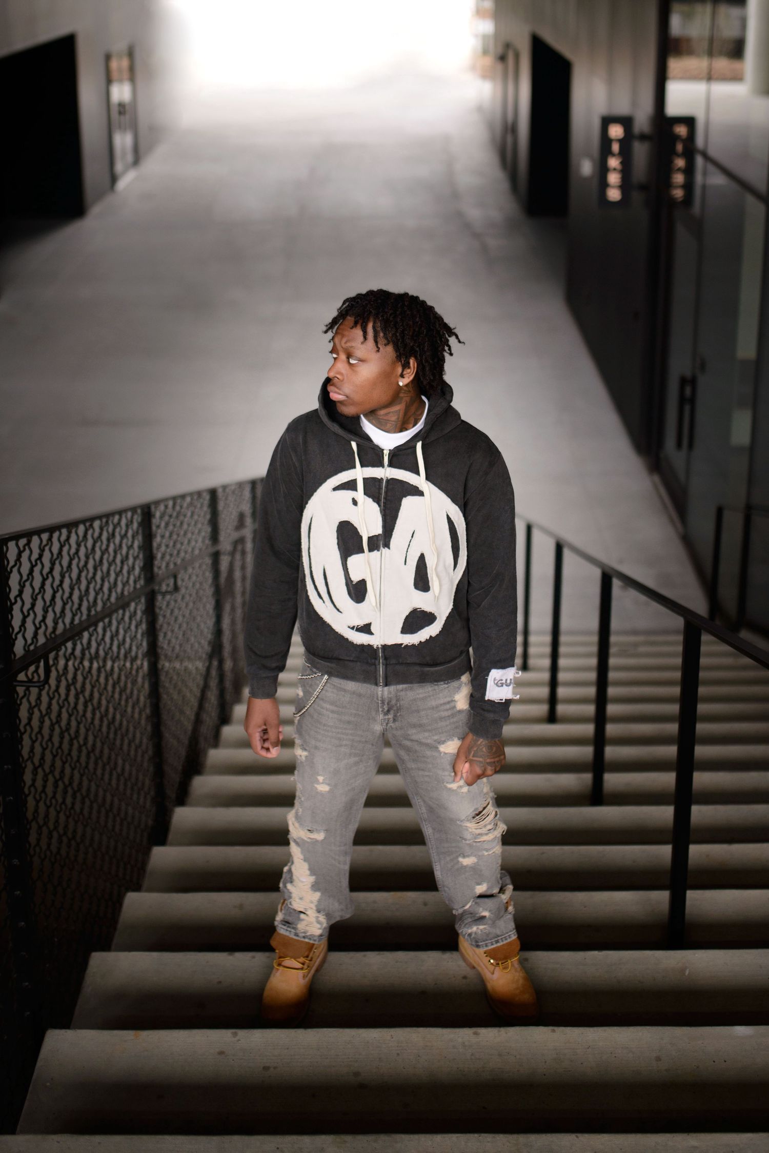 Young man in a dark GA graphic hoodie and ripped jeans stands on outdoor stairs wearing tan boots.