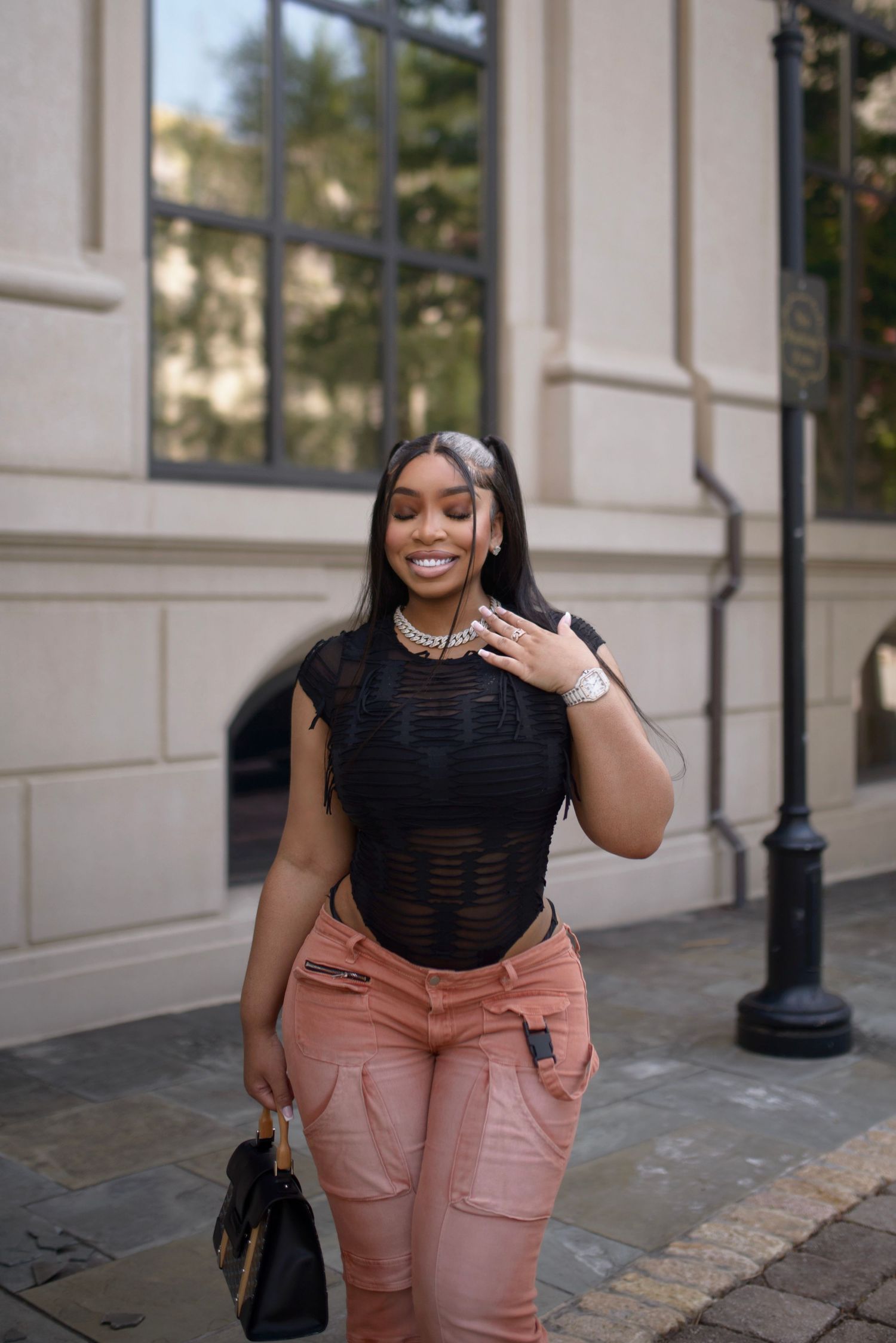 Smiling woman in black mesh top and pink cargo pants walks outdoors carrying a designer handbag.