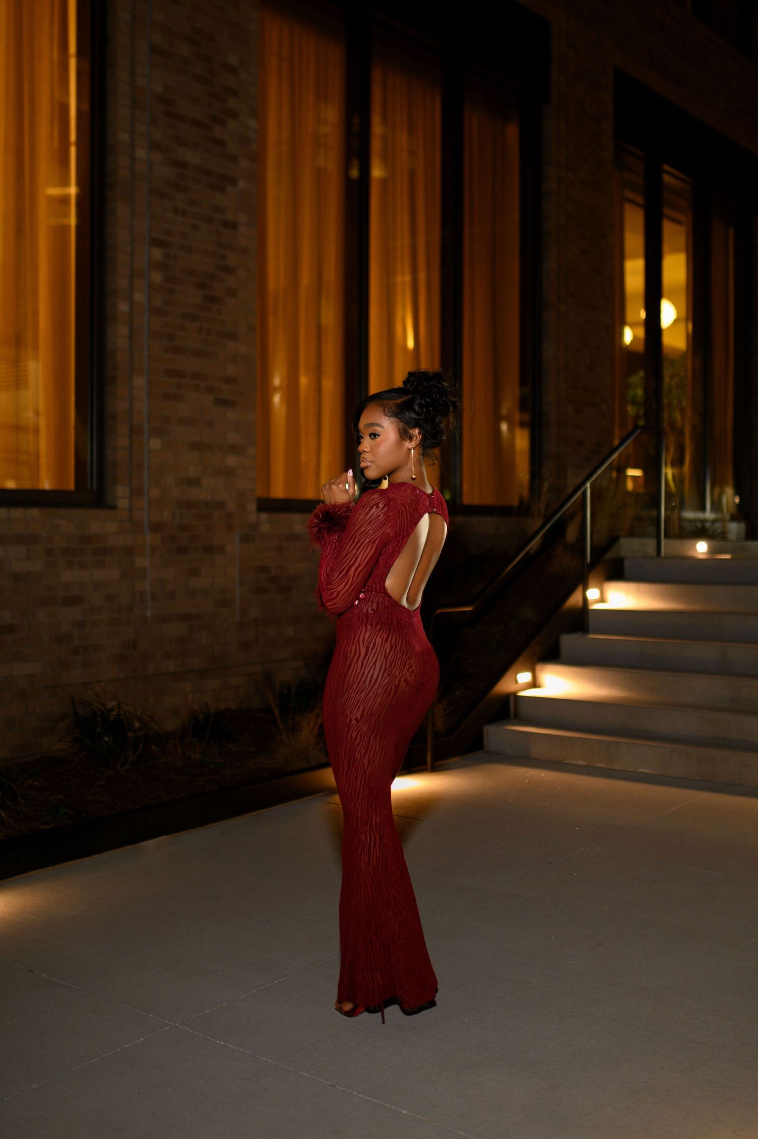 Woman in a red sequin backless gown poses on a lit outdoor stairway at night.