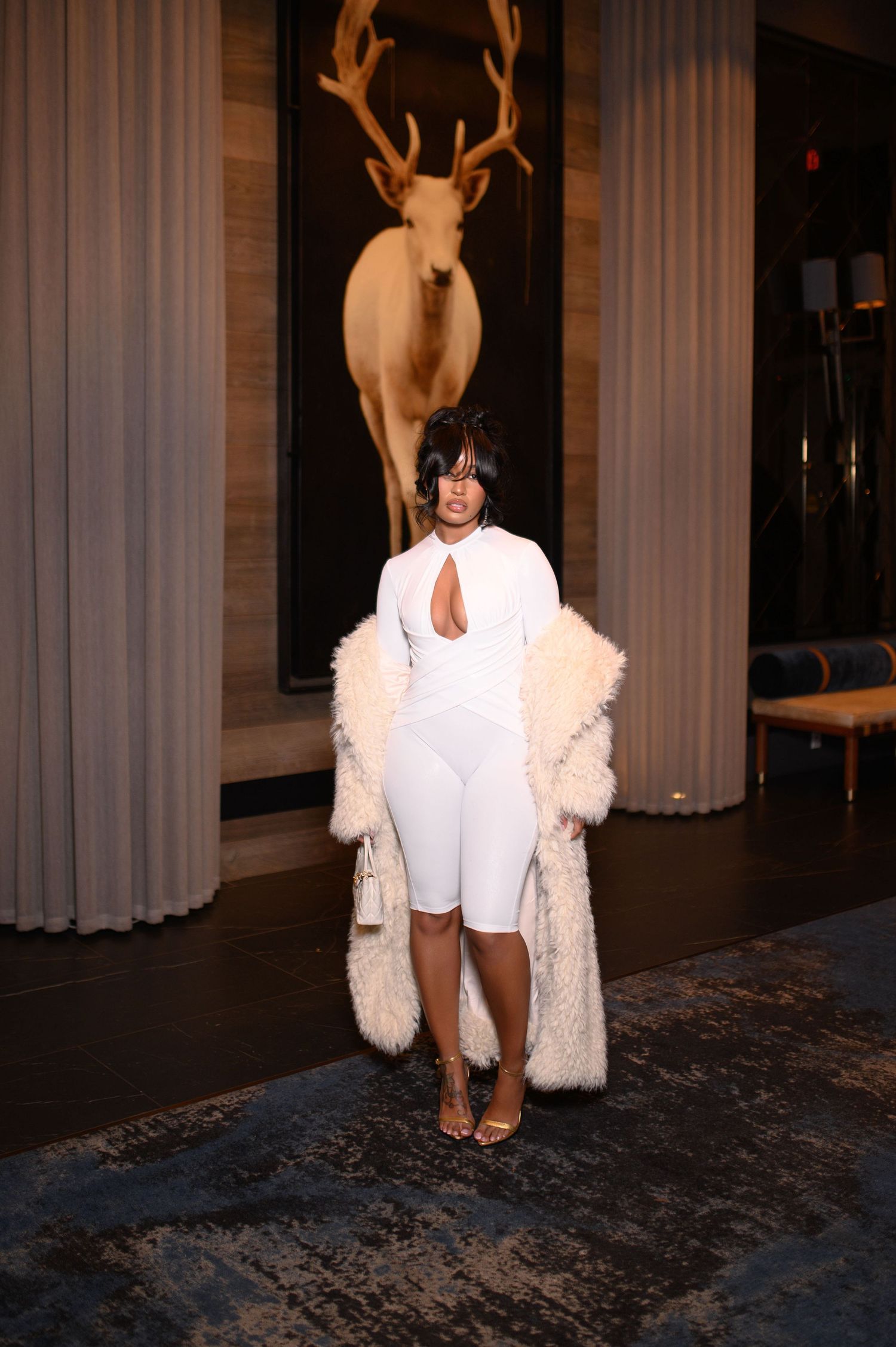 Woman in white cutout bodysuit and fluffy cream coat poses before deer artwork.