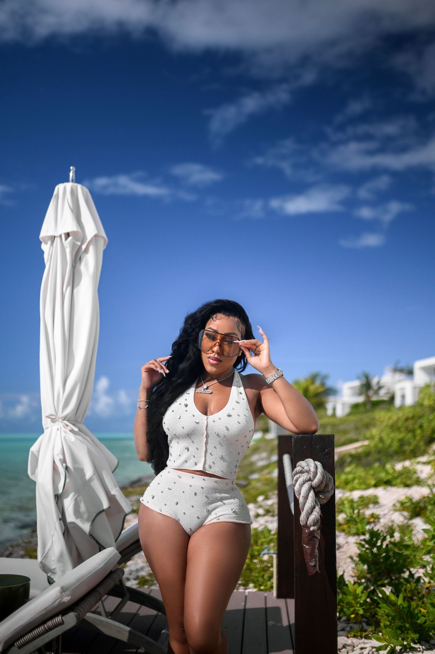 Curvy woman in white crop top and shorts adjusting sunglasses by beach lounge chair.
