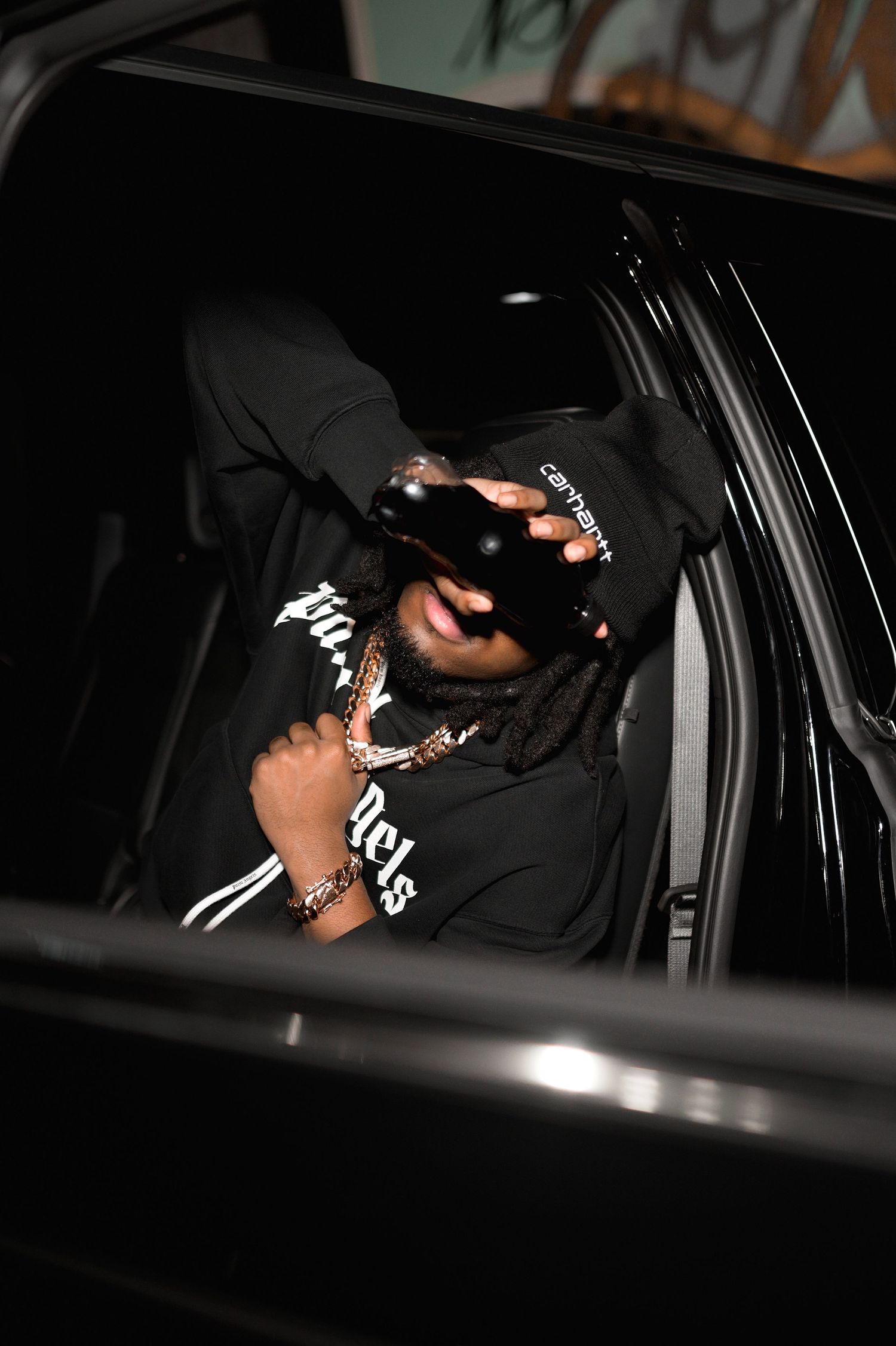 Man in black Carhartt hoodie covering face with hand while sitting in a black vehicle at night.