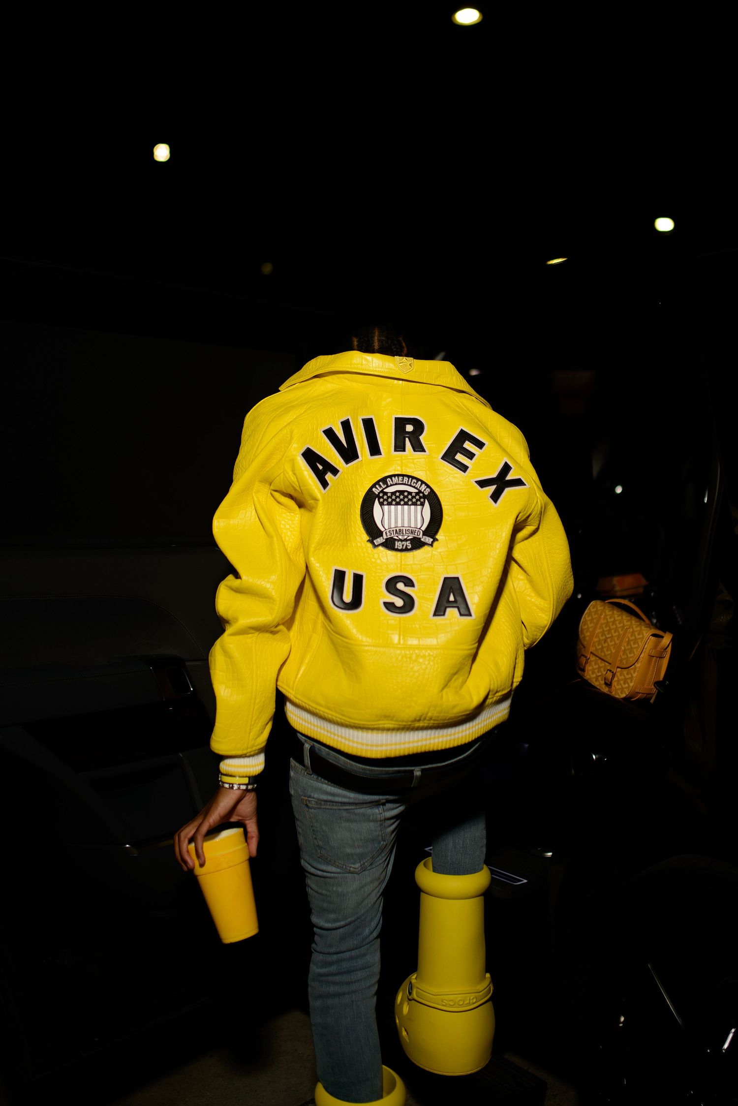 Person wearing a bright yellow Avirex USA varsity jacket and jeans photographed from behind at night.