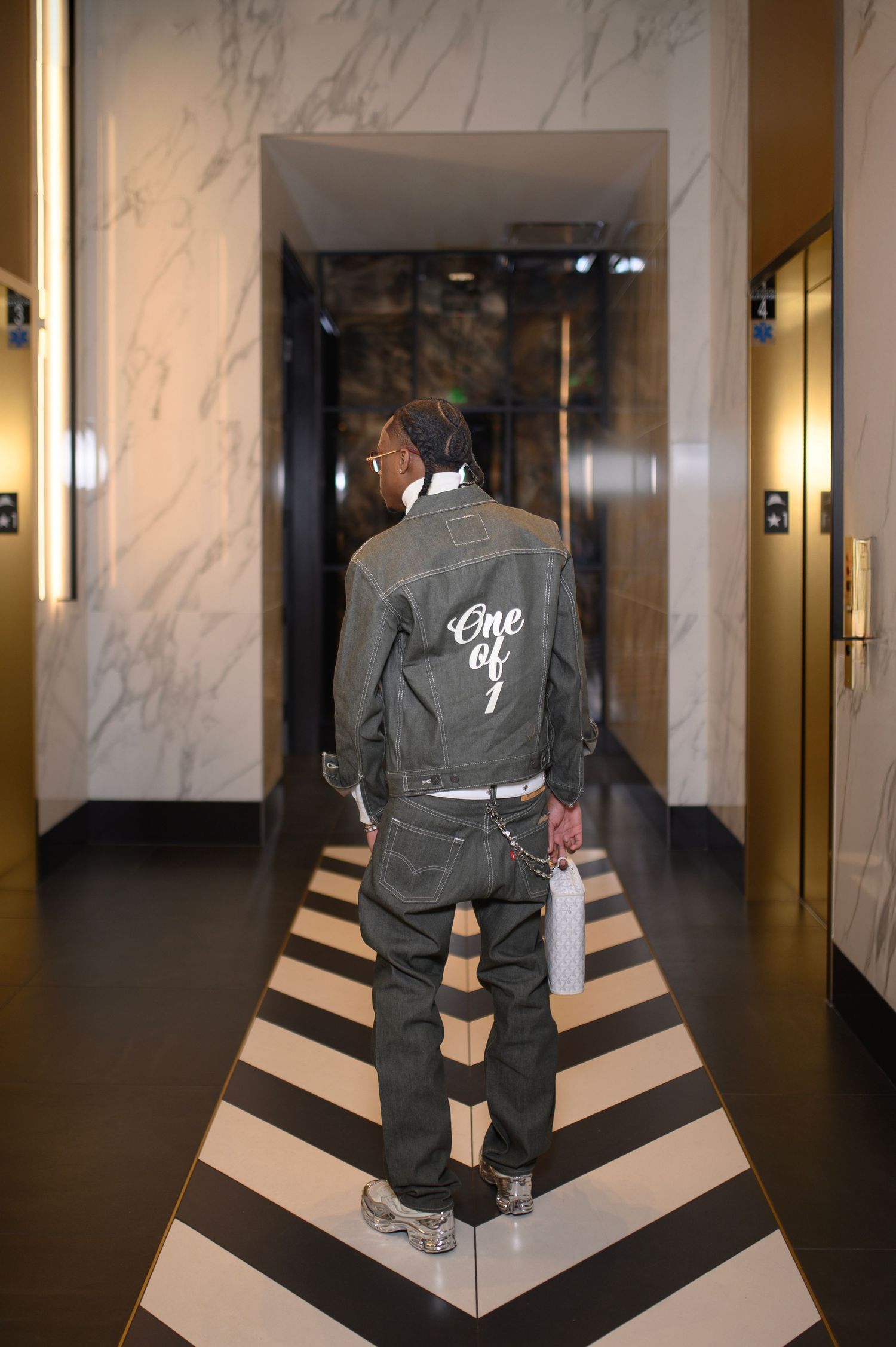 Man in denim jacket with 'One of 1' print walks through luxury marble hotel corridor.