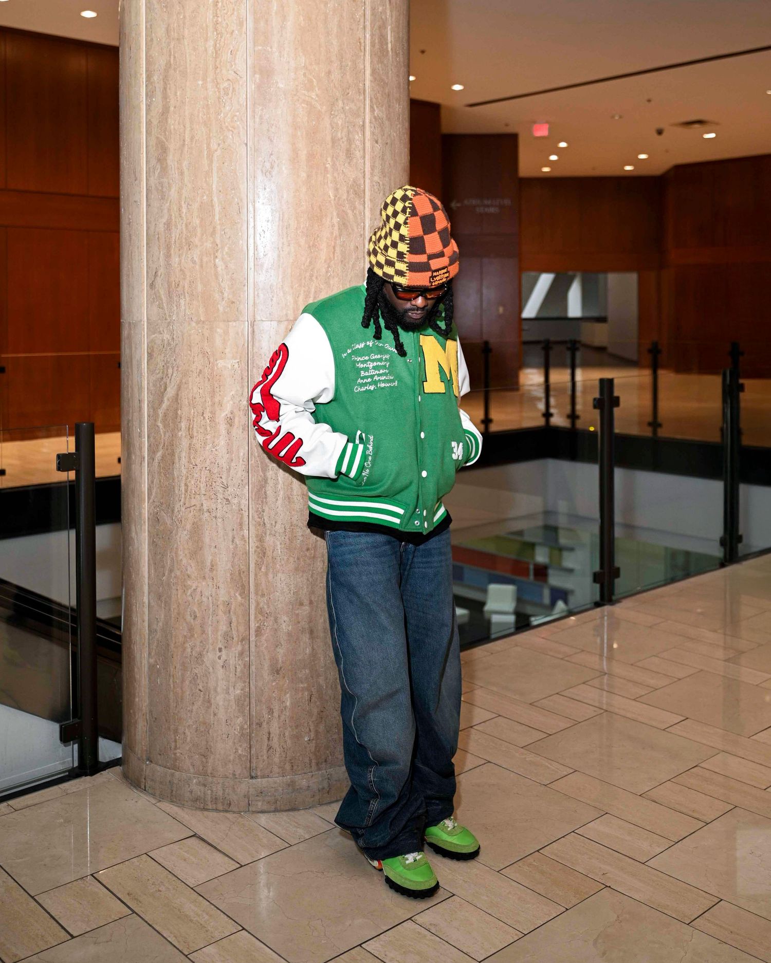 Man in green varsity jacket and checkered beanie leaning on lobby column.