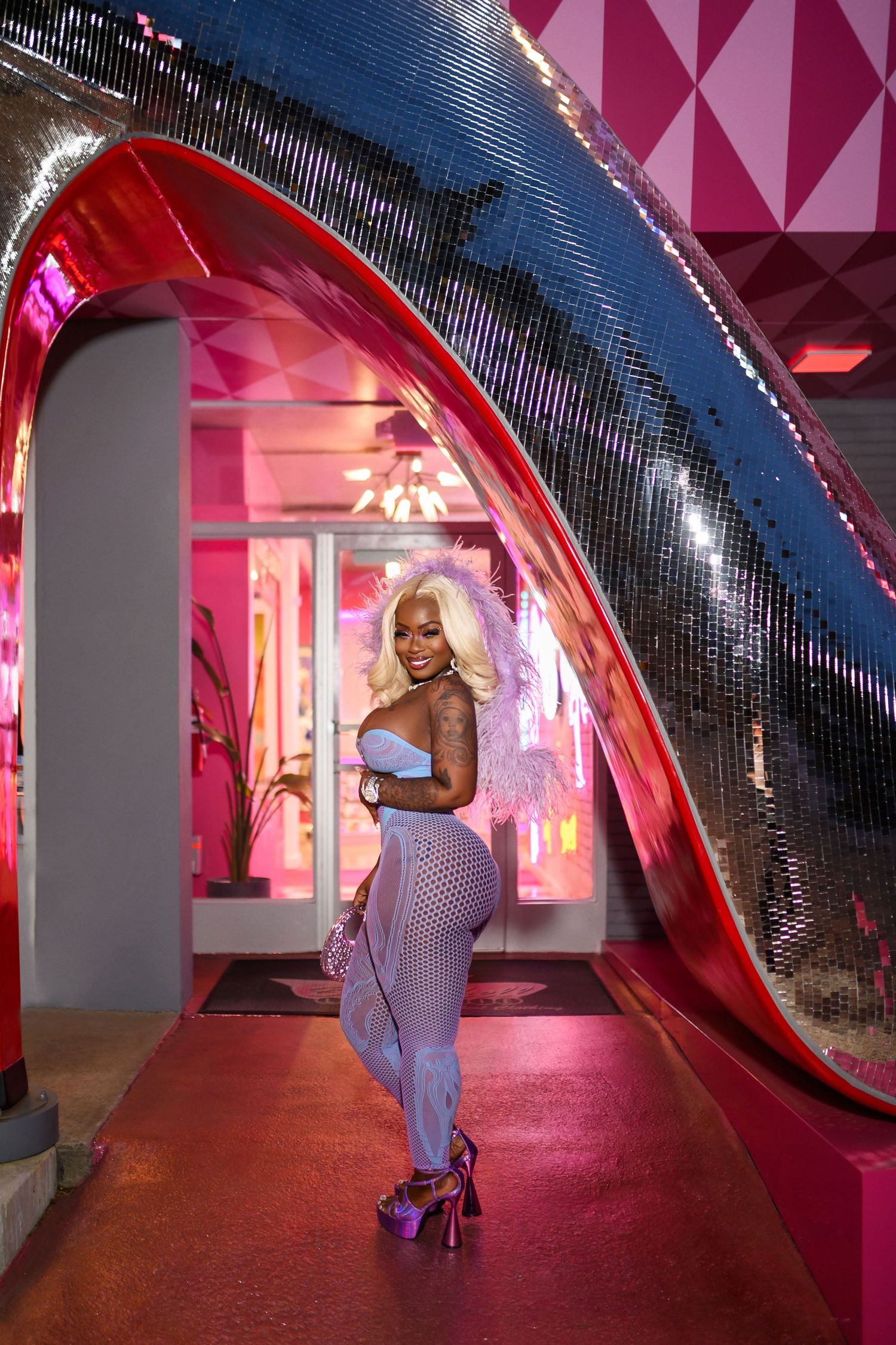 Woman in blue fishnet outfit with platinum blonde hair posing under disco ball arch in pink venue.