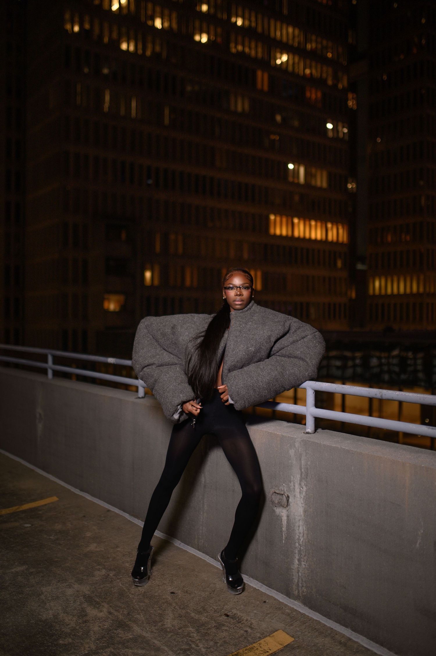 Model in oversized gray puffer jacket and black tights poses on urban rooftop at night.