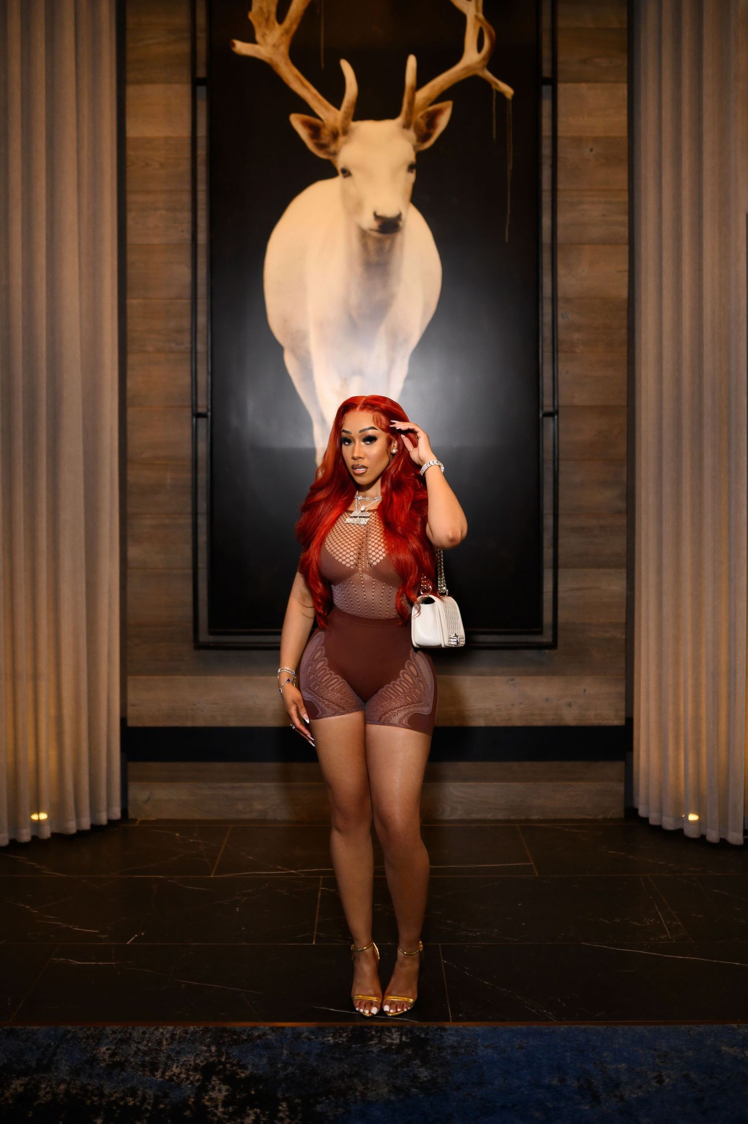 Woman with red hair in brown mesh romper standing beneath mounted deer antlers in elegant interior.