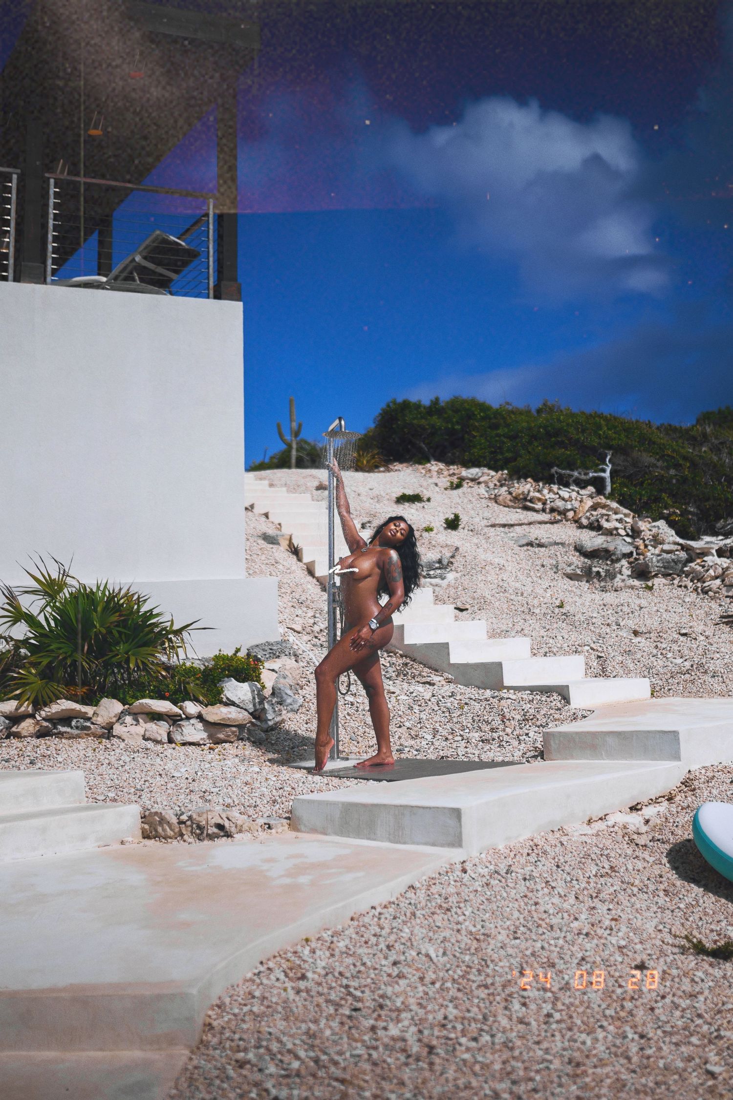 Woman posing outdoors near white stone steps and a pole with a rocky hillside and blue sky background.