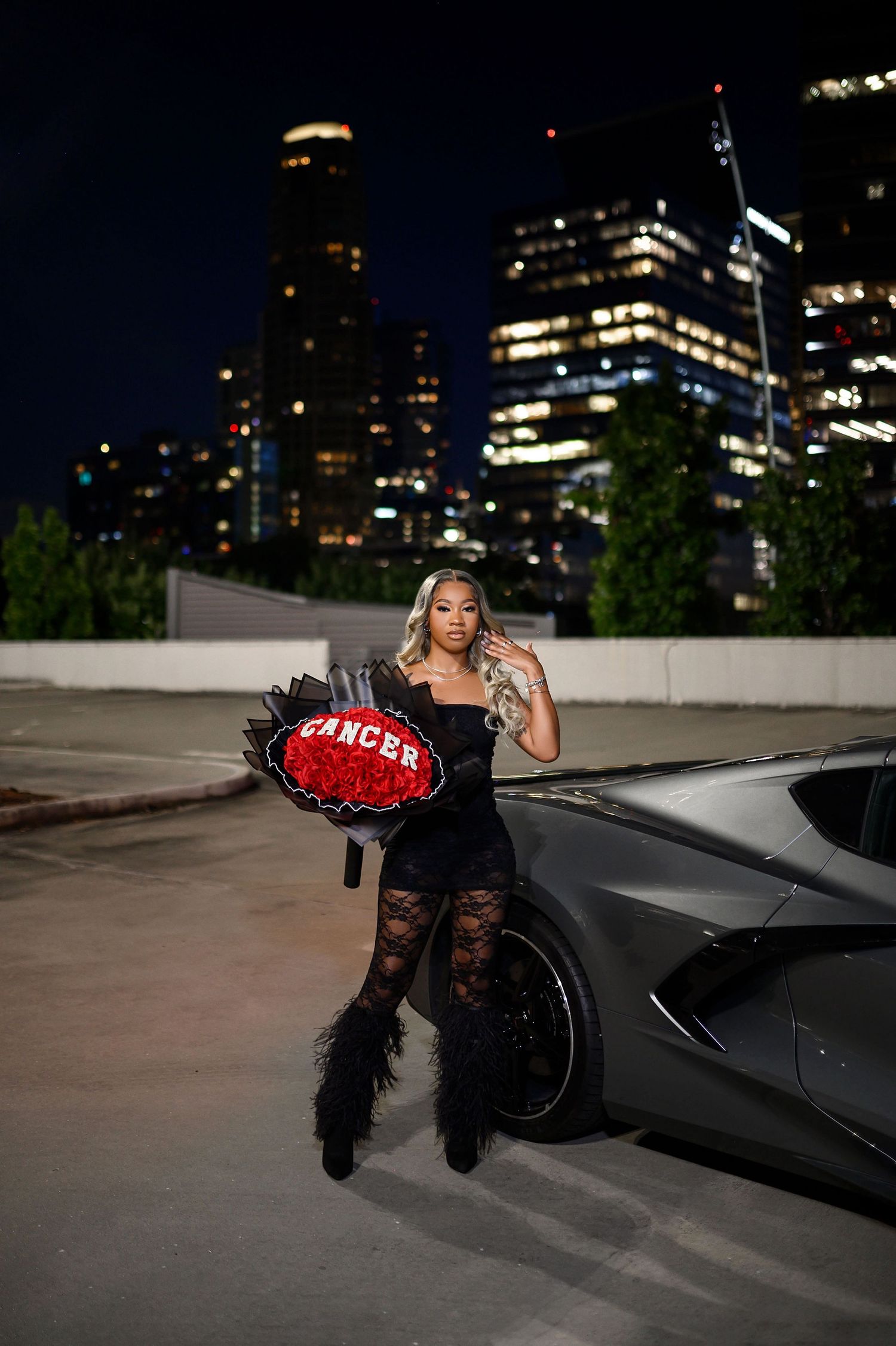 Blonde woman in black dress holding Cancer rose bouquet leaning on sports car with city skyline.